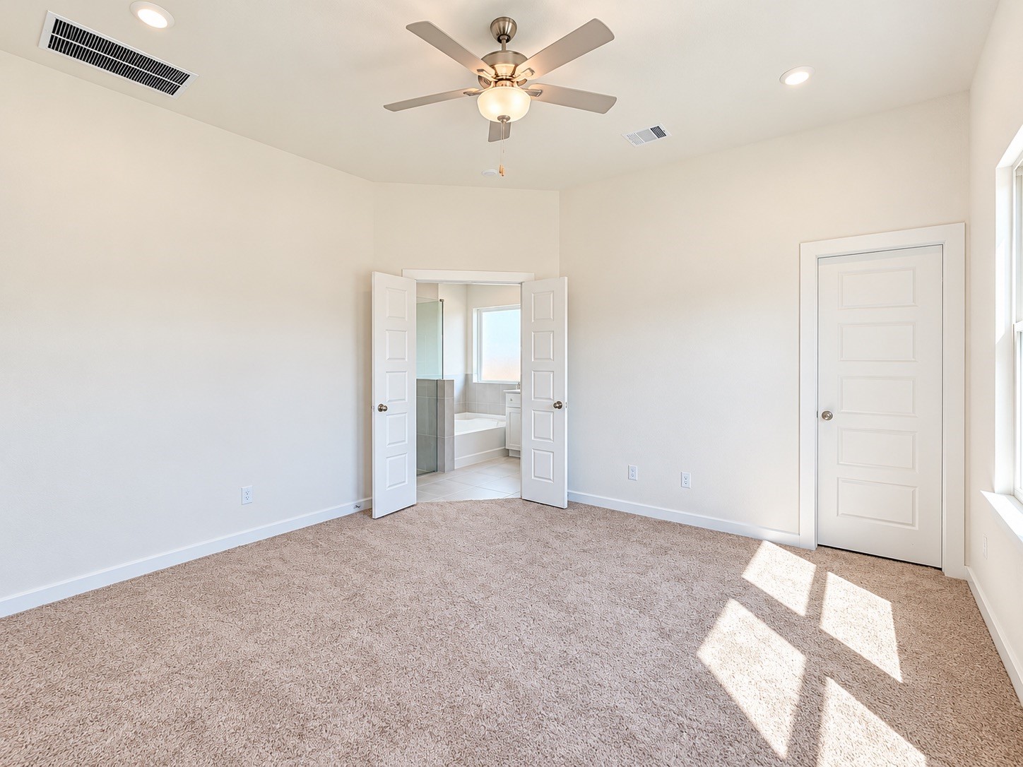 Spacious primary bedroom with plush carpeting, modern ceiling fan, and ample natural light, featuring double doors leading to an en-suite bathroom.