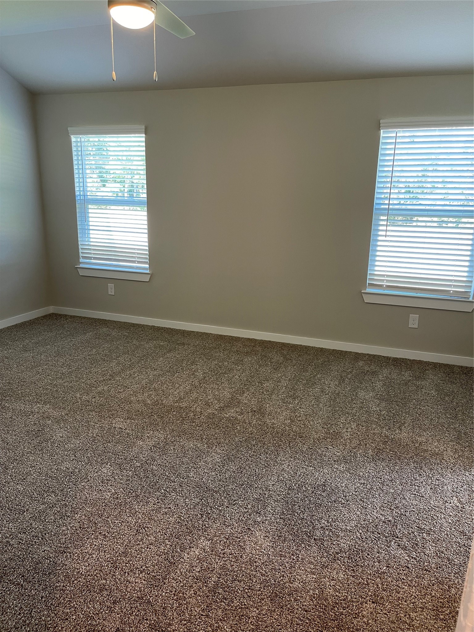 Spacious carpeted room with two large windows offering ample natural light and a ceiling fan for comfort. Spacious carpeted room with two large windows offering ample natural light and a ceiling fan for comfort.