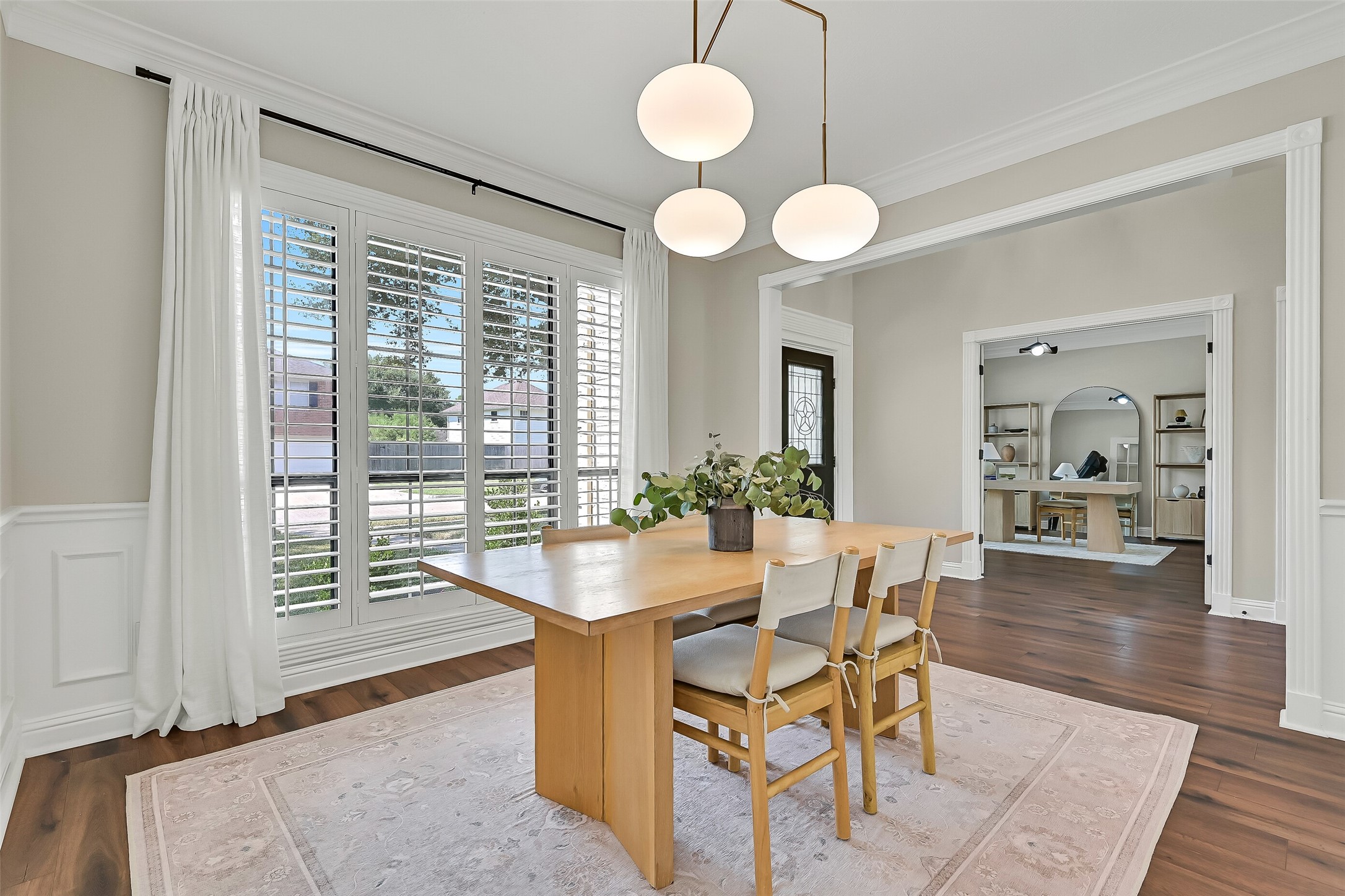 Another view of the formal dining room highlights its generous proportions and easy connection to the foyer. Detailed molding, plantation shutters, a contemporary chandelier & warm luxury vinyl plank flooring create an inviting space for gatherings.