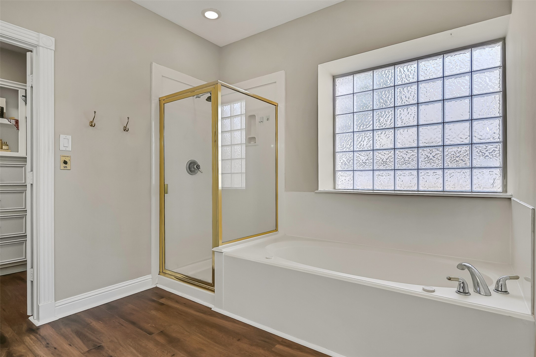 Opposite the vanities, a jetted soaking tub and separate glass-enclosed shower complete the primary bath. A large glass-block window fills the space with natural light while maintaining privacy, and warm LVP flooring carries throughout.