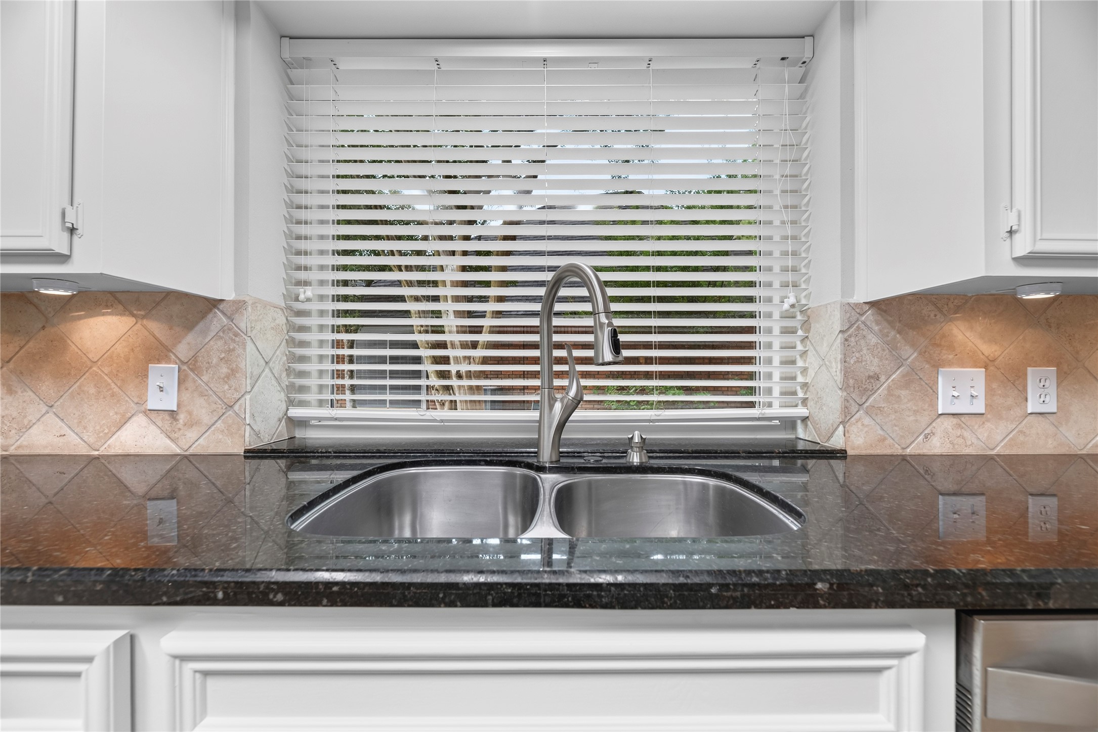 A double-basin stainless steel sink, sleek granite countertops, tumbled stone backsplash, under-cabinet lighting, an updated window above the sink & bright overhead lighting create a practical workspace for daily kitchen tasks.