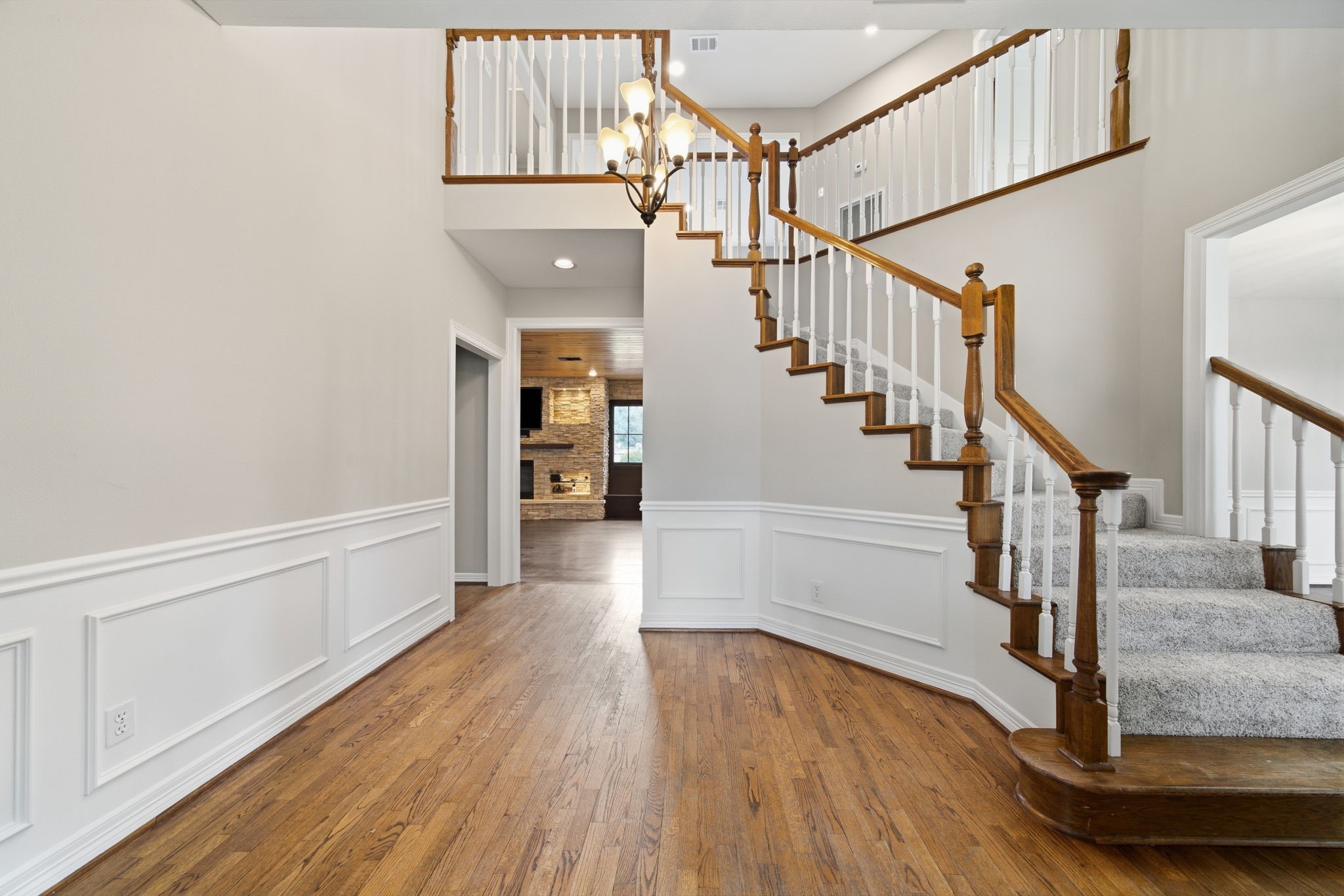 Elegant foyer with rich hardwood floors and a grand staircase, leading to a cozy living area adorned with a brick fireplace. Large storage closet under staircase with shelving!