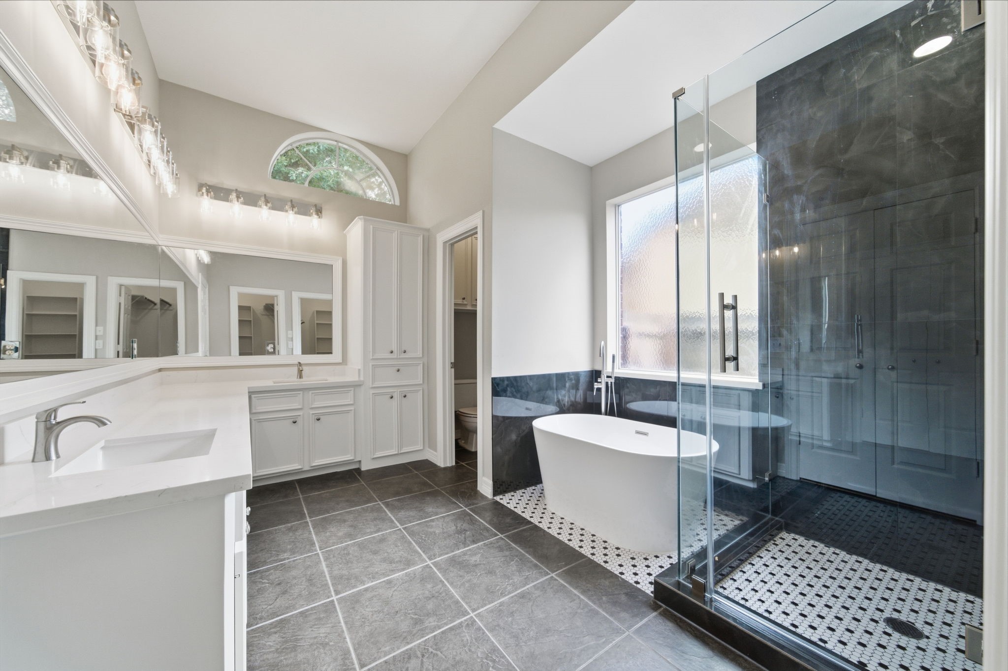 Renovated luxurious bathroom featuring a sleek freestanding bathtub, spacious glass-enclosed shower, dual vanities with elegant lighting, and abundant natural light from a large window.