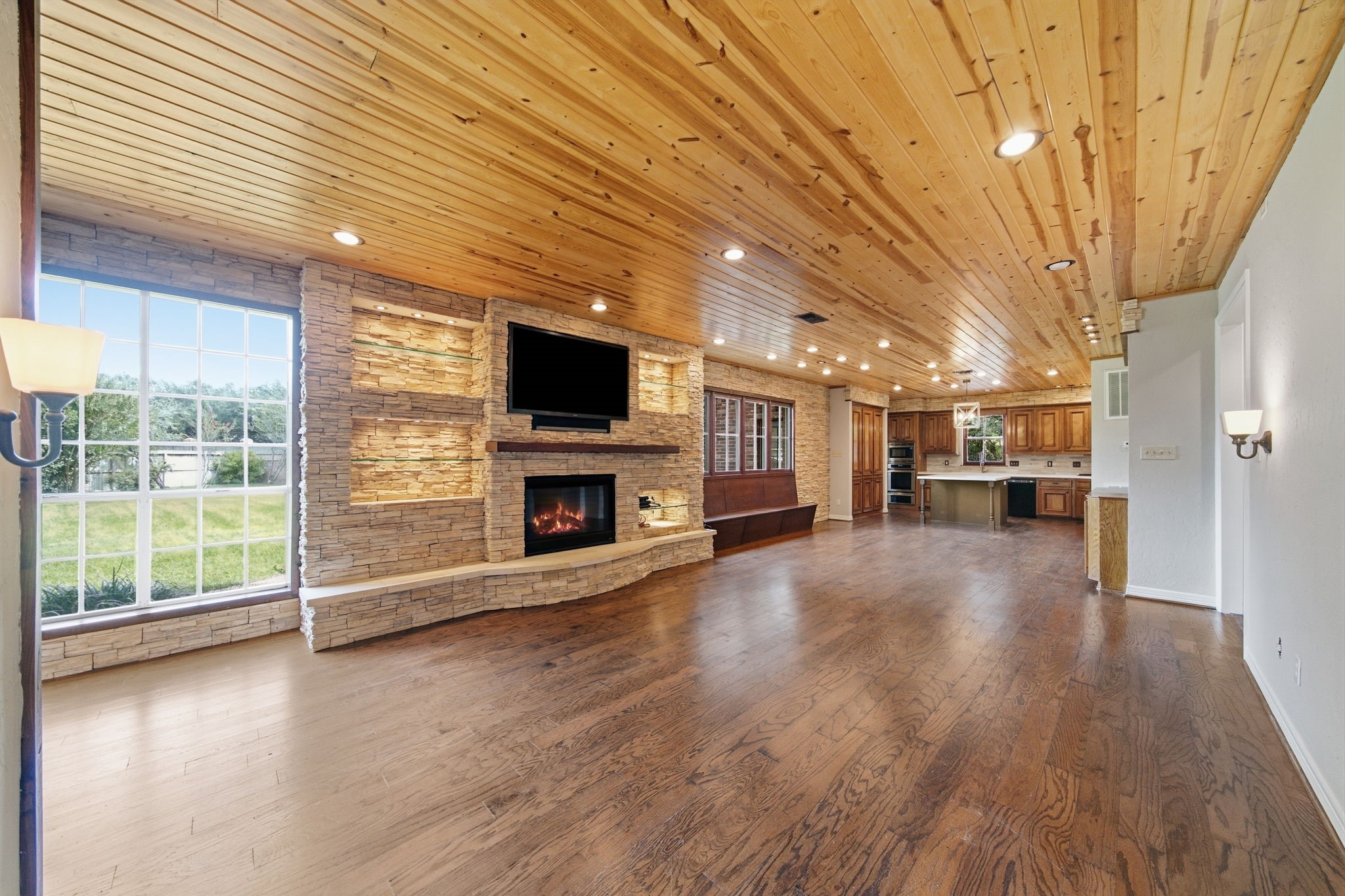 Spacious open-concept living area featuring a cozy fireplace with stone accents, rich wood flooring, and a warm wood ceiling, seamlessly flowing into the modern kitchen. Expansive windows provide abundant natural light and scenic views.