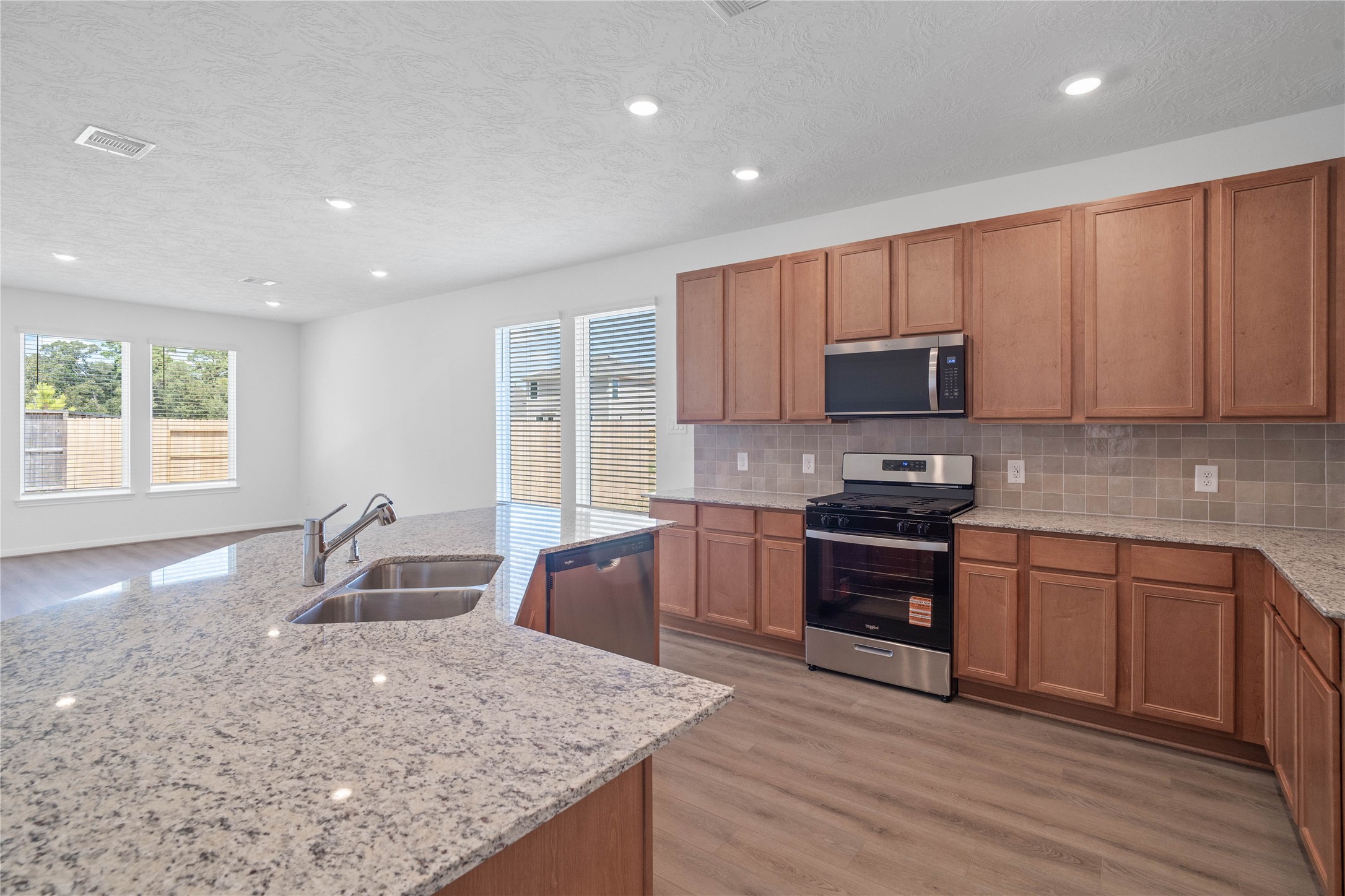 This spacious kitchen boasts high ceilings, Woodmont Cody cabinets in a Toffee stain, Granite White Napoli countertops, stainless steel Whirlpool appliances, a modern Daltile backsplash, recessed lighting, windows with blinds, an island with added breakfast bar, and a pantryall overlooking the expansive Great Room.