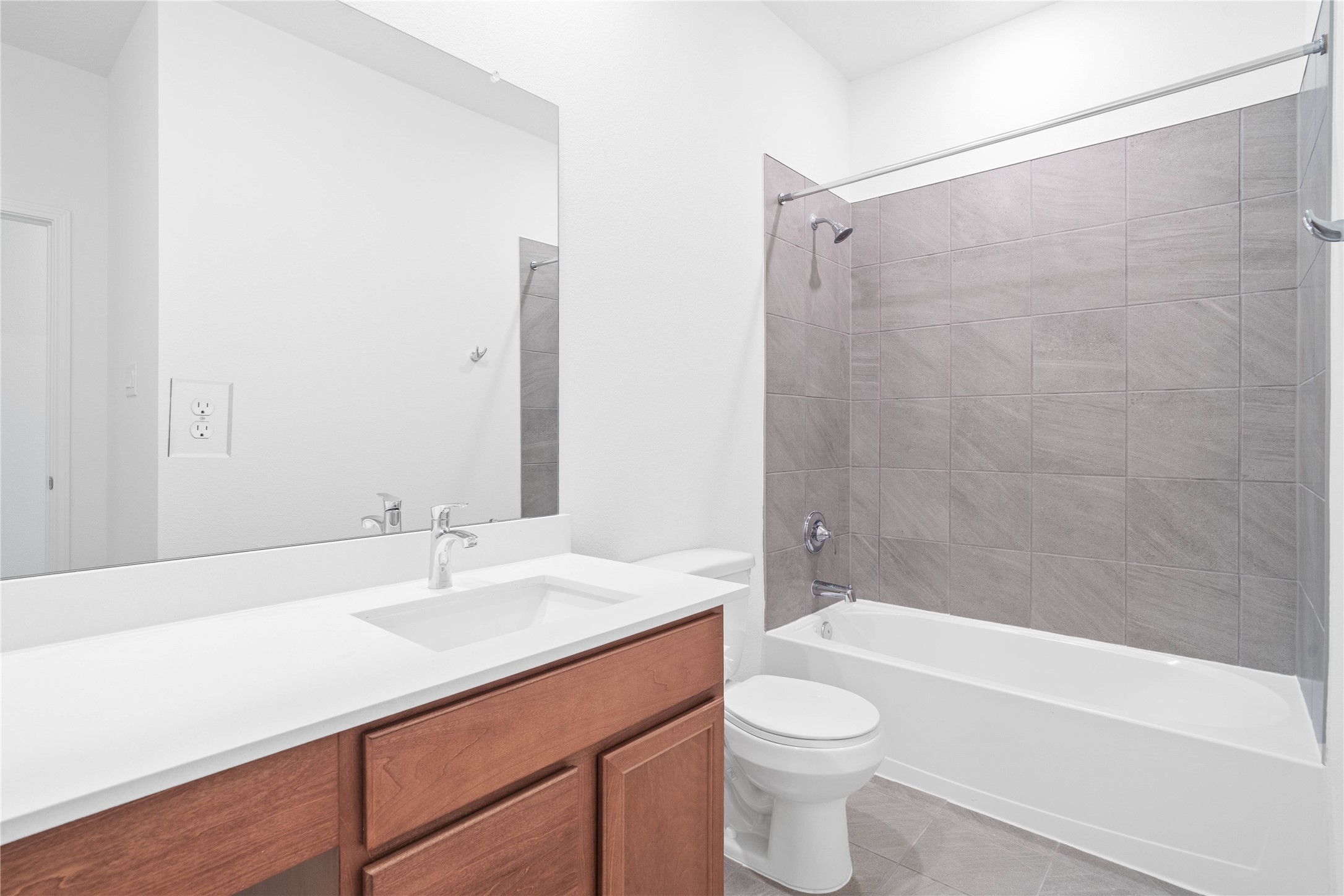 The secondary bathroom offers tile flooring, a bath/shower combination with a stylish tile surround, rich stained wood cabinetry with added knee space and elegant light countertops, a mirror, and sleek fixtures with modern finishes.