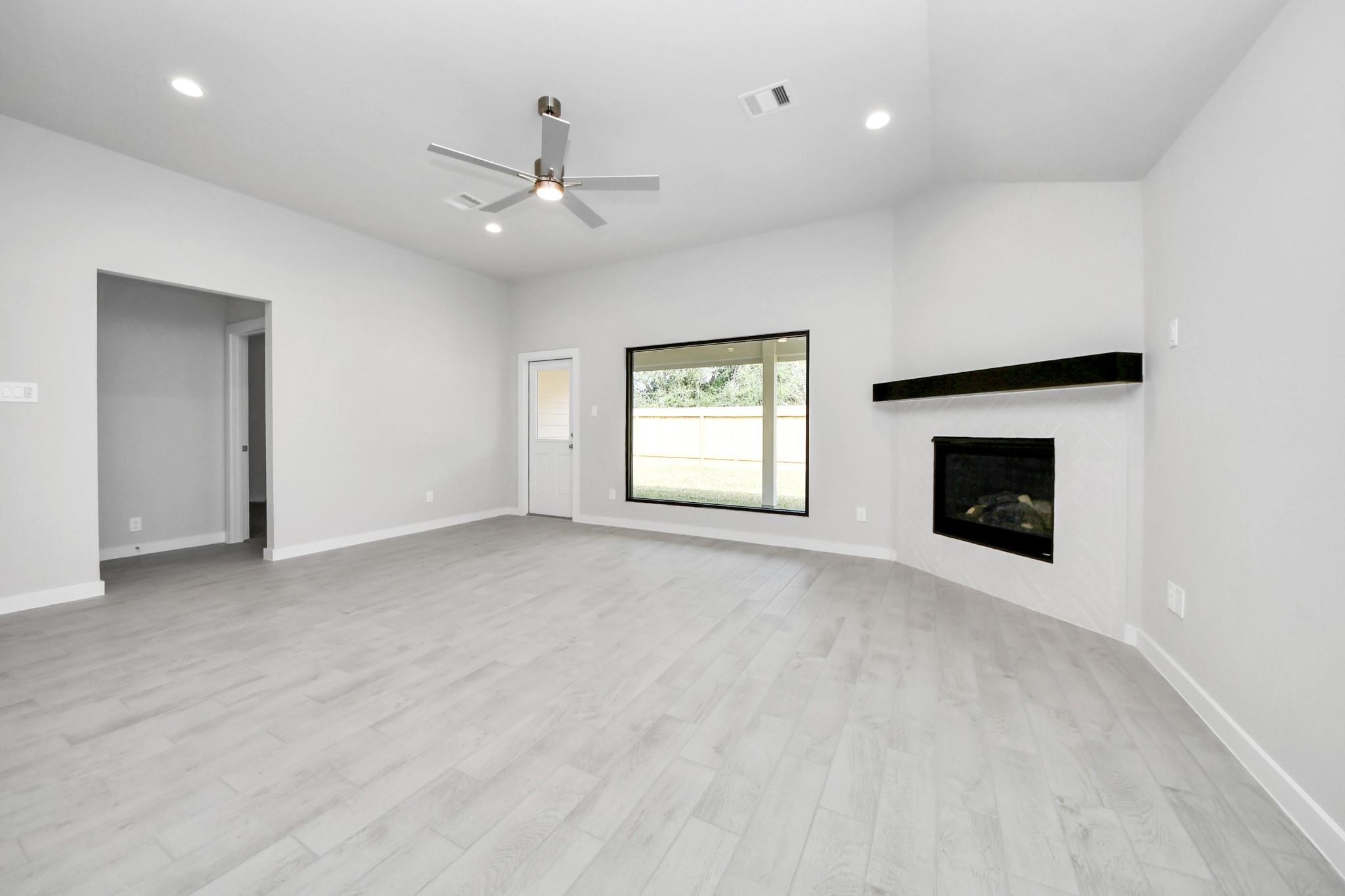 Spacious living room featuring sleek wood flooring, a contemporary ceiling fan, and a cozy corner fireplace, complemented by large windows for abundant natural light.