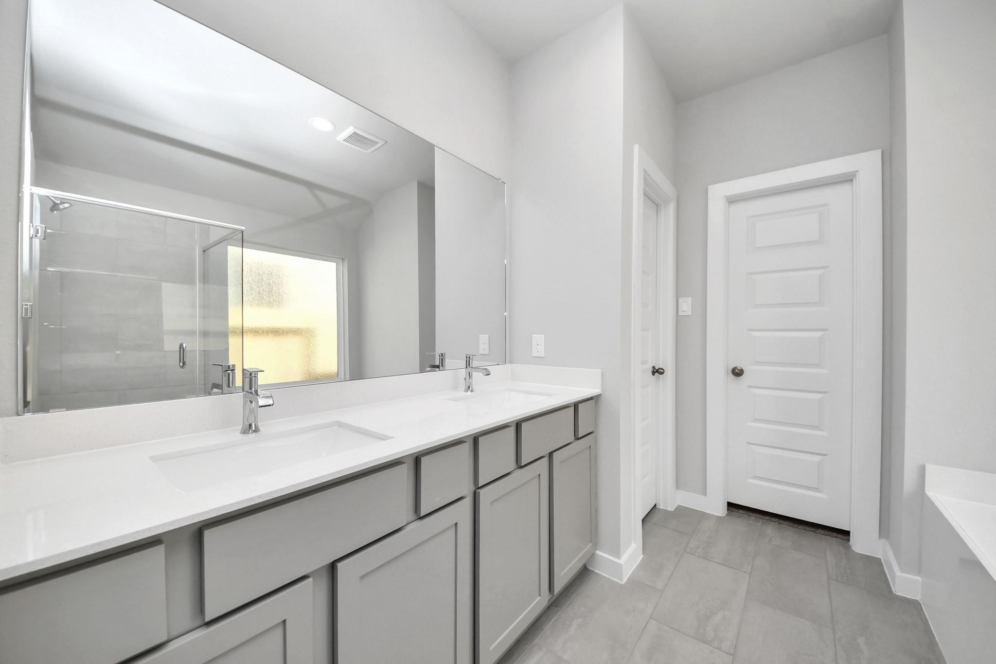 Spacious bathroom featuring a double vanity with sleek gray cabinetry, a large mirror, and modern fixtures, complemented by a glass-enclosed shower and elegant tile flooring.