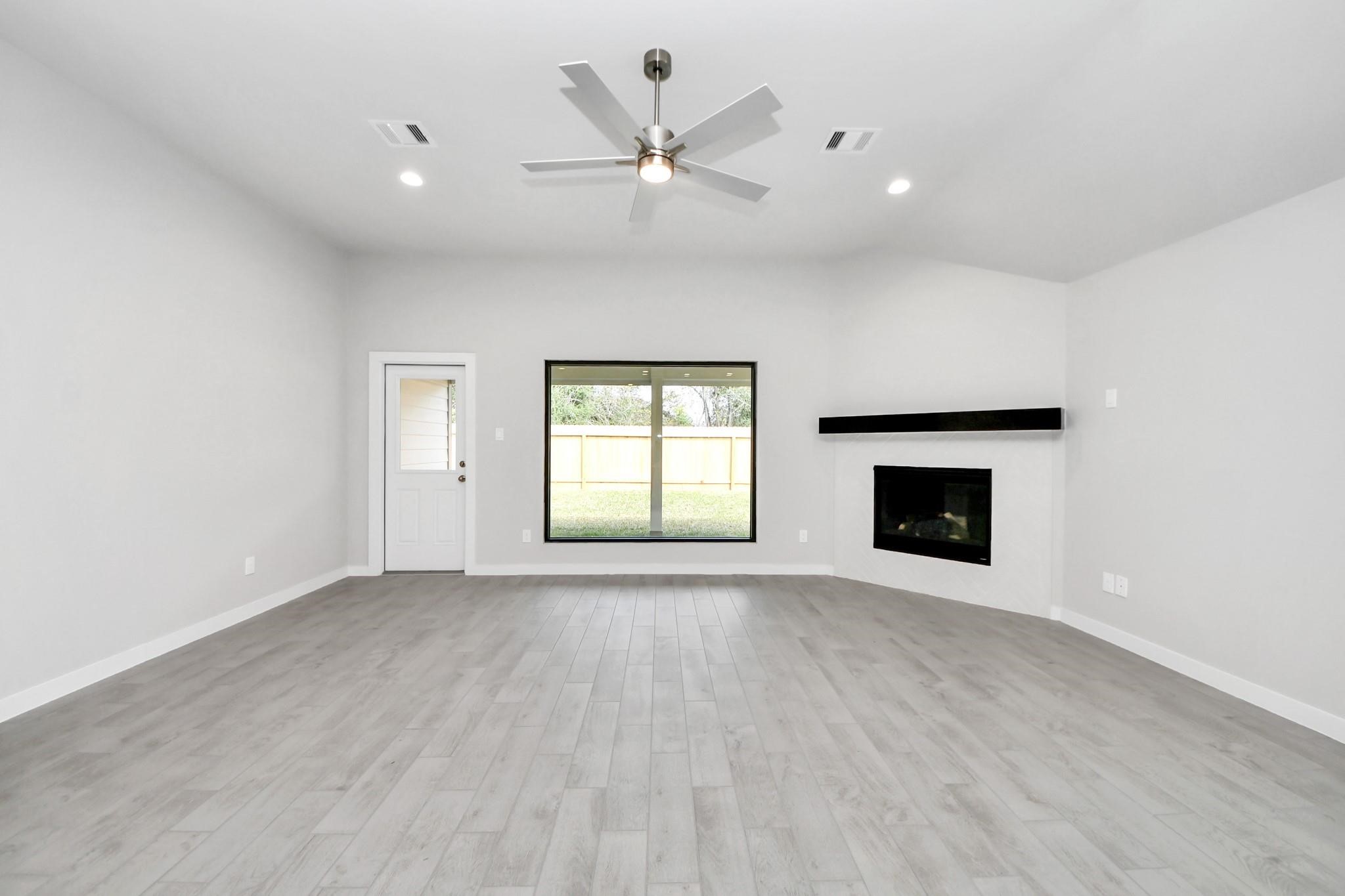 Spacious living room with sleek hardwood floors, a modern ceiling fan, and a cozy corner fireplace, all enhanced by ample natural light from a large window and convenient patio access. Spacious living room with sleek hardwood floors, a modern ceiling fan, and a cozy corner fireplace, all enhanced by ample natural light from a large window and convenient patio access.