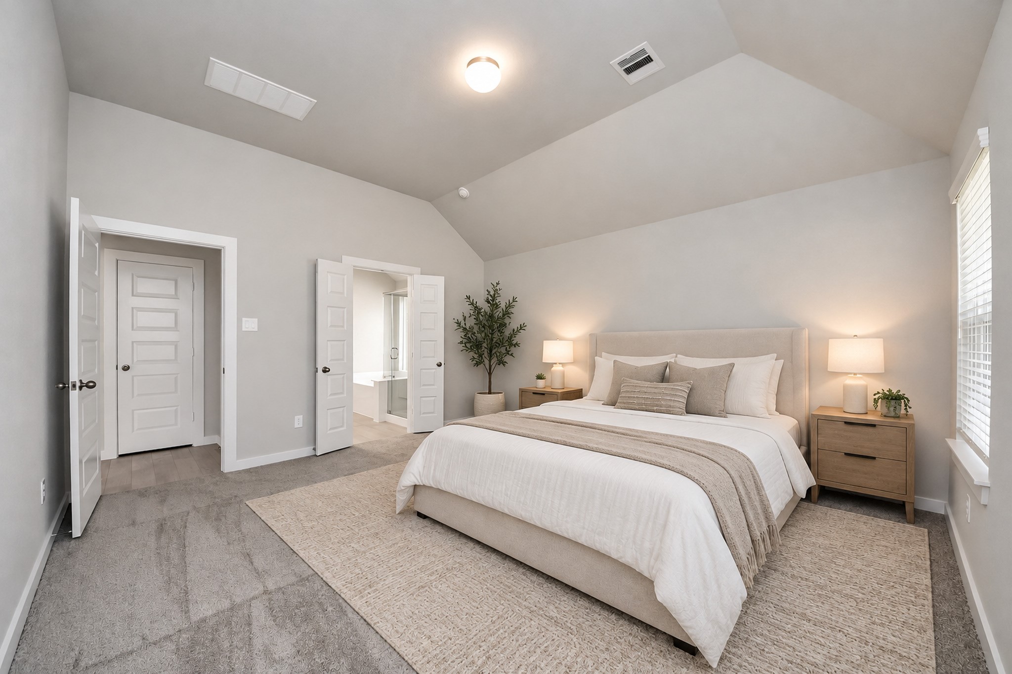 Spacious bedroom with vaulted ceiling, plush carpeting, and soft neutral tones. Features a cozy bed, stylish nightstands, and ample natural light. Double doors lead to an en-suite bathroom for added convenience.