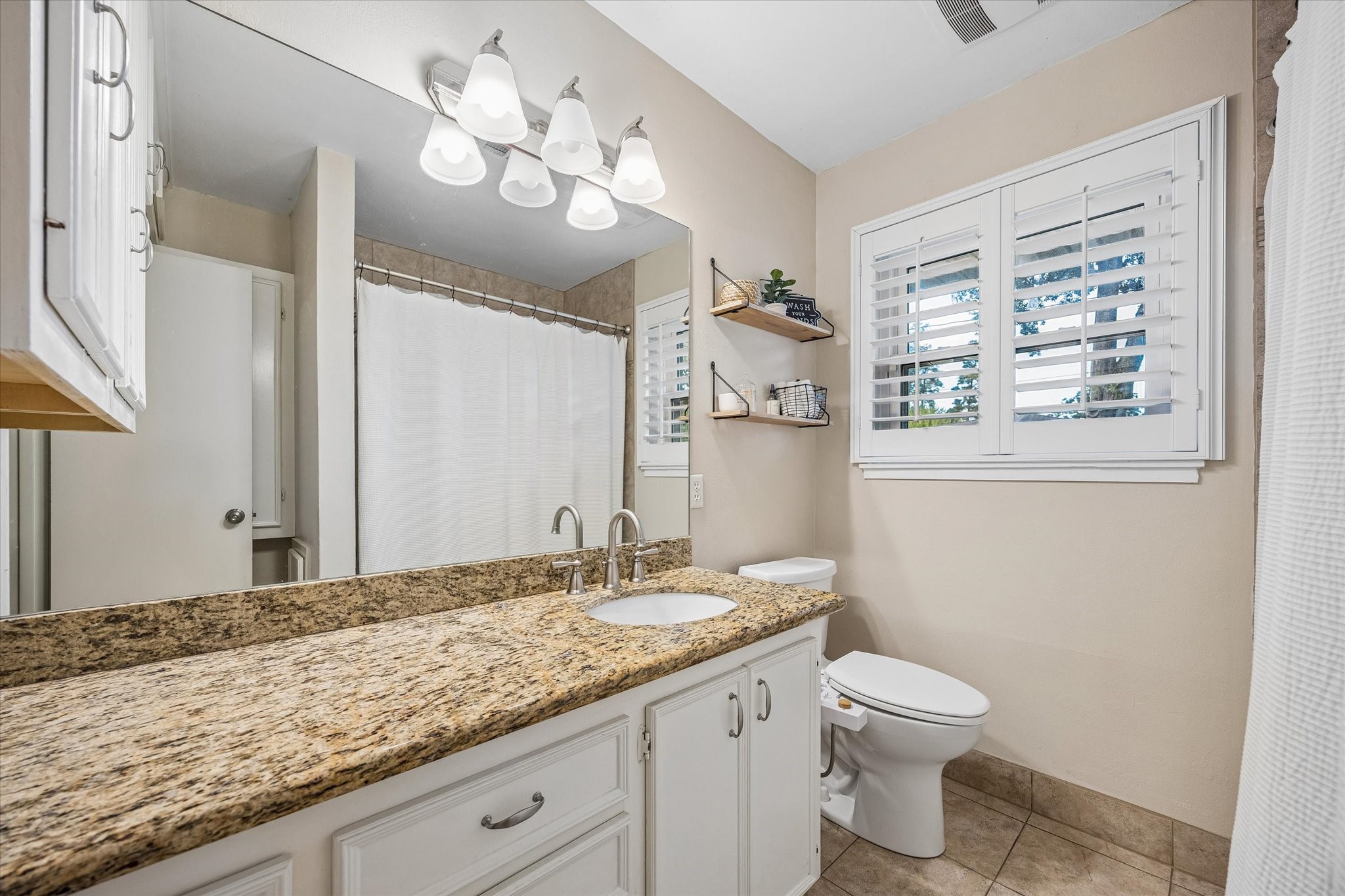 Wide vanity counter provides room for daily routines, with cabinet storage below to keep essentials organized. A shuttered window brings natural light and privacy control to the bathroom near the bedrooms.