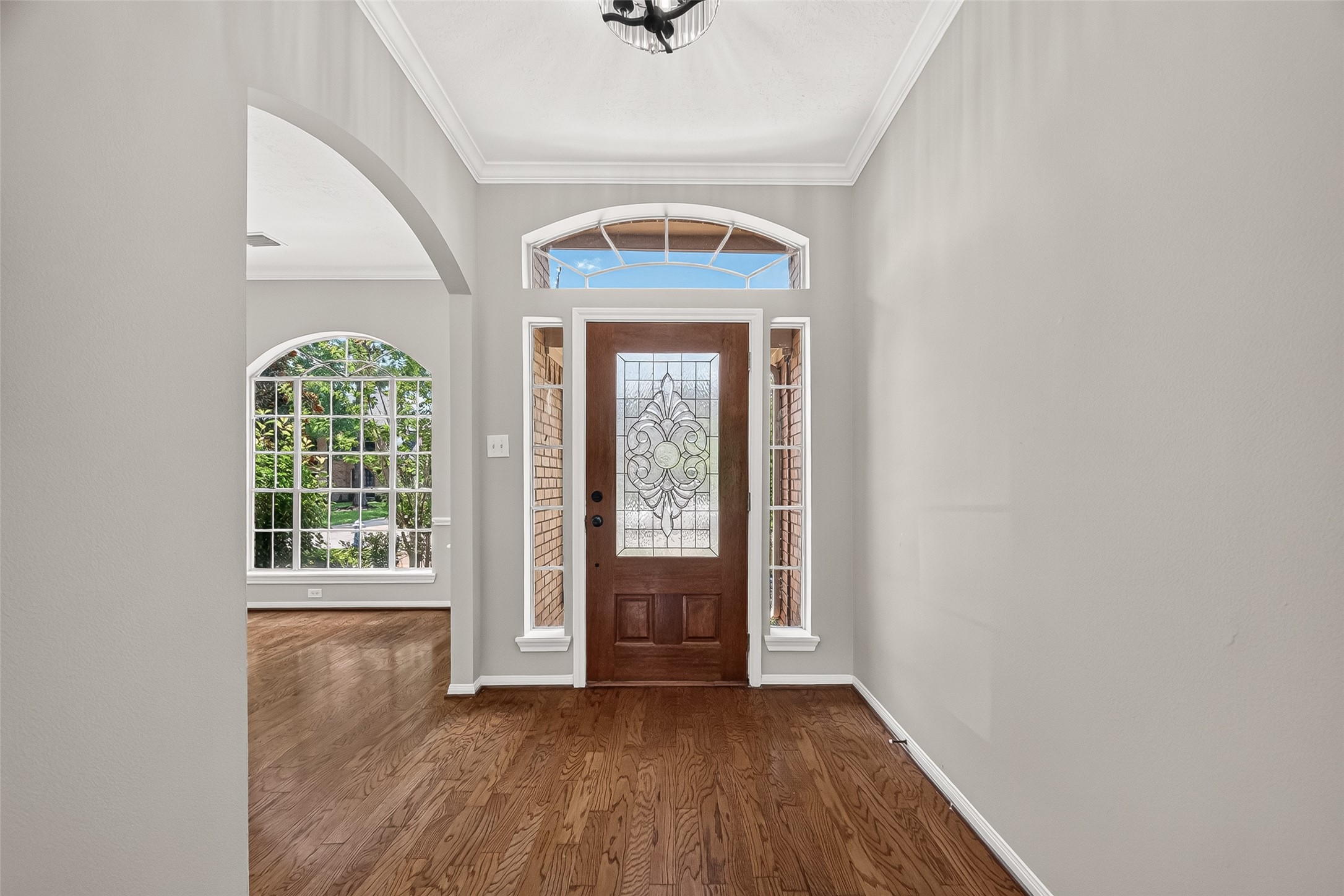 Entryway with a beautifully detailed wooden door and sidelights, complemented by rich hardwood flooring and abundant natural light from large arched windows.