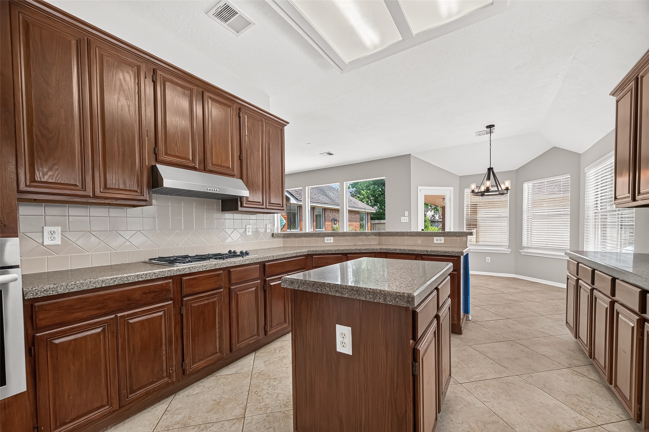Spacious kitchen with rich wood cabinetry, granite countertops, and a central island. Ample natural light fills the room, highlighting the tile flooring and open layout, perfect for gatherings.