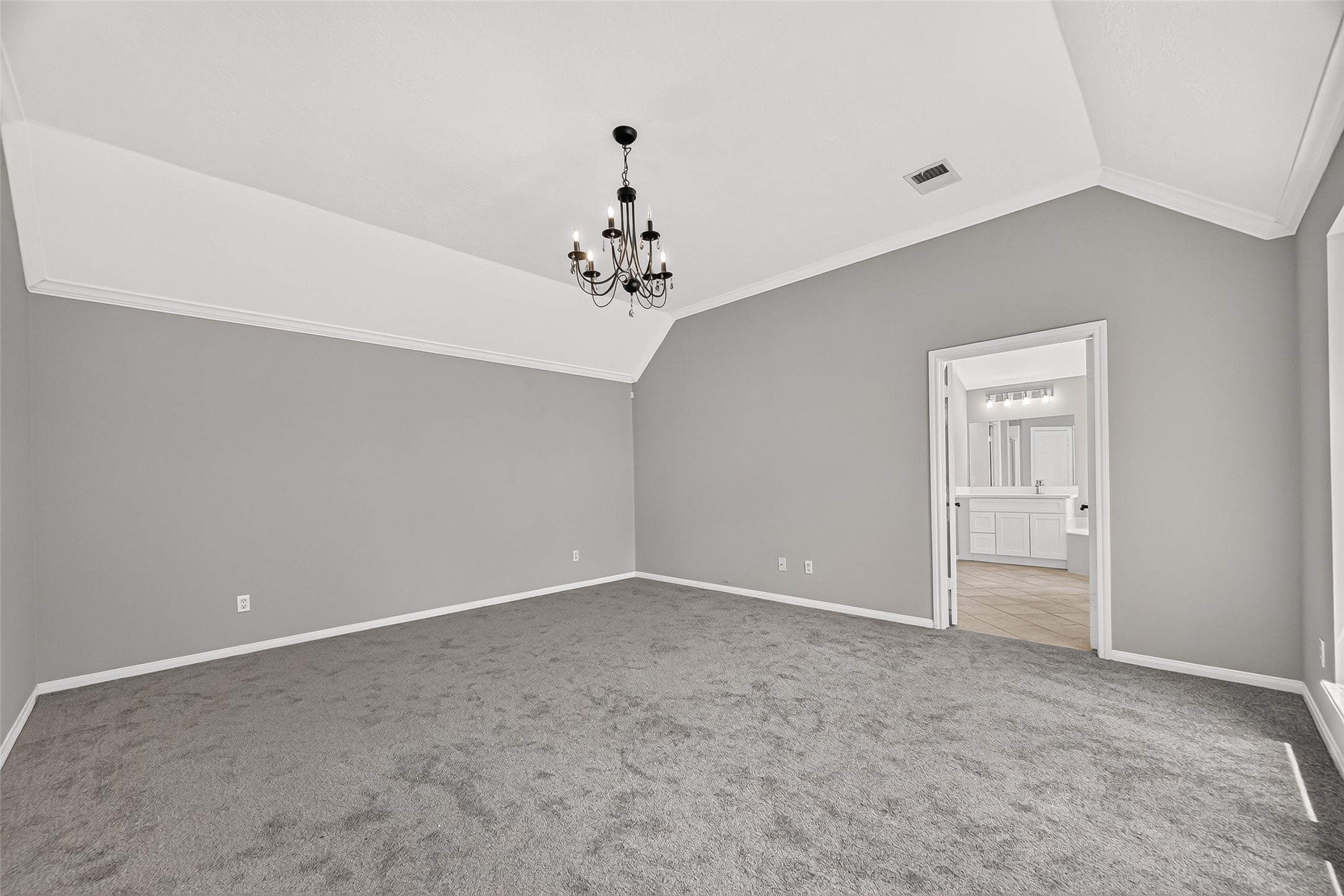 Spacious bedroom featuring elegant vaulted ceilings, a chic chandelier, and plush carpeting, leading to a bright en-suite bathroom for added privacy and convenience.