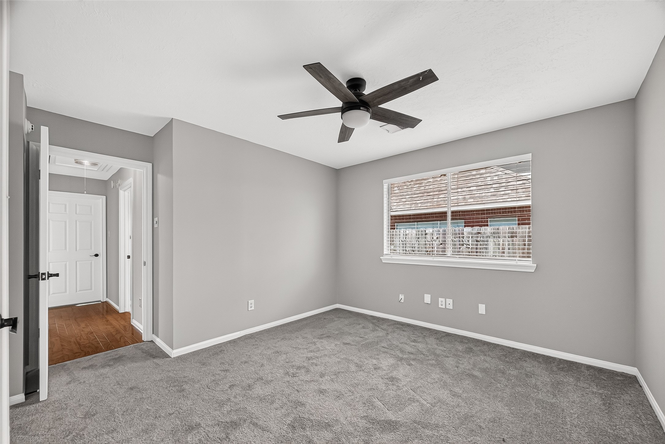 Spacious bedroom with neutral gray walls, plush carpet, and a modern ceiling fan, complemented by a large window for natural light.