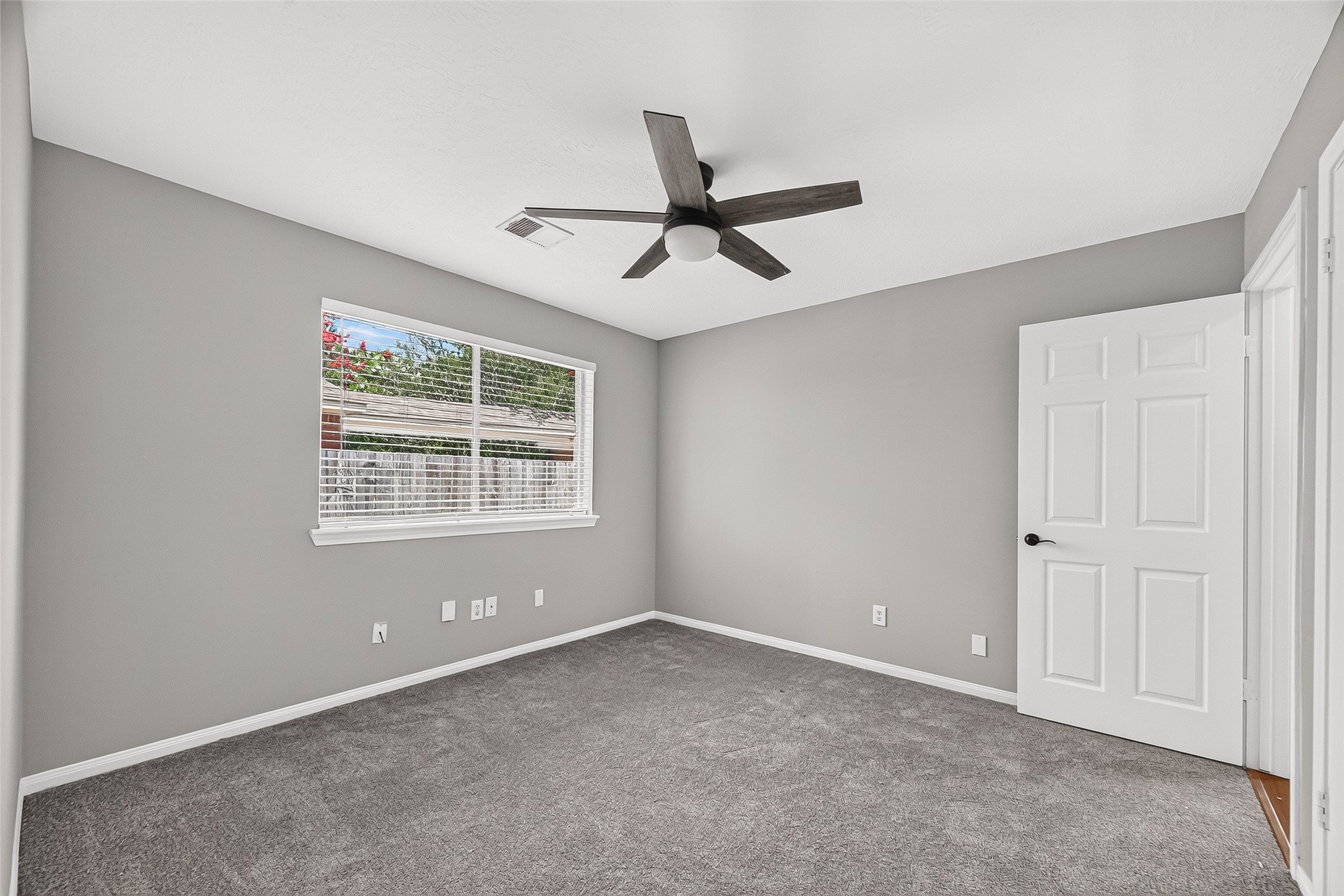 Bright and inviting bedroom featuring fresh gray walls, plush carpeting, and a modern ceiling fan, with ample natural light streaming through a large window.