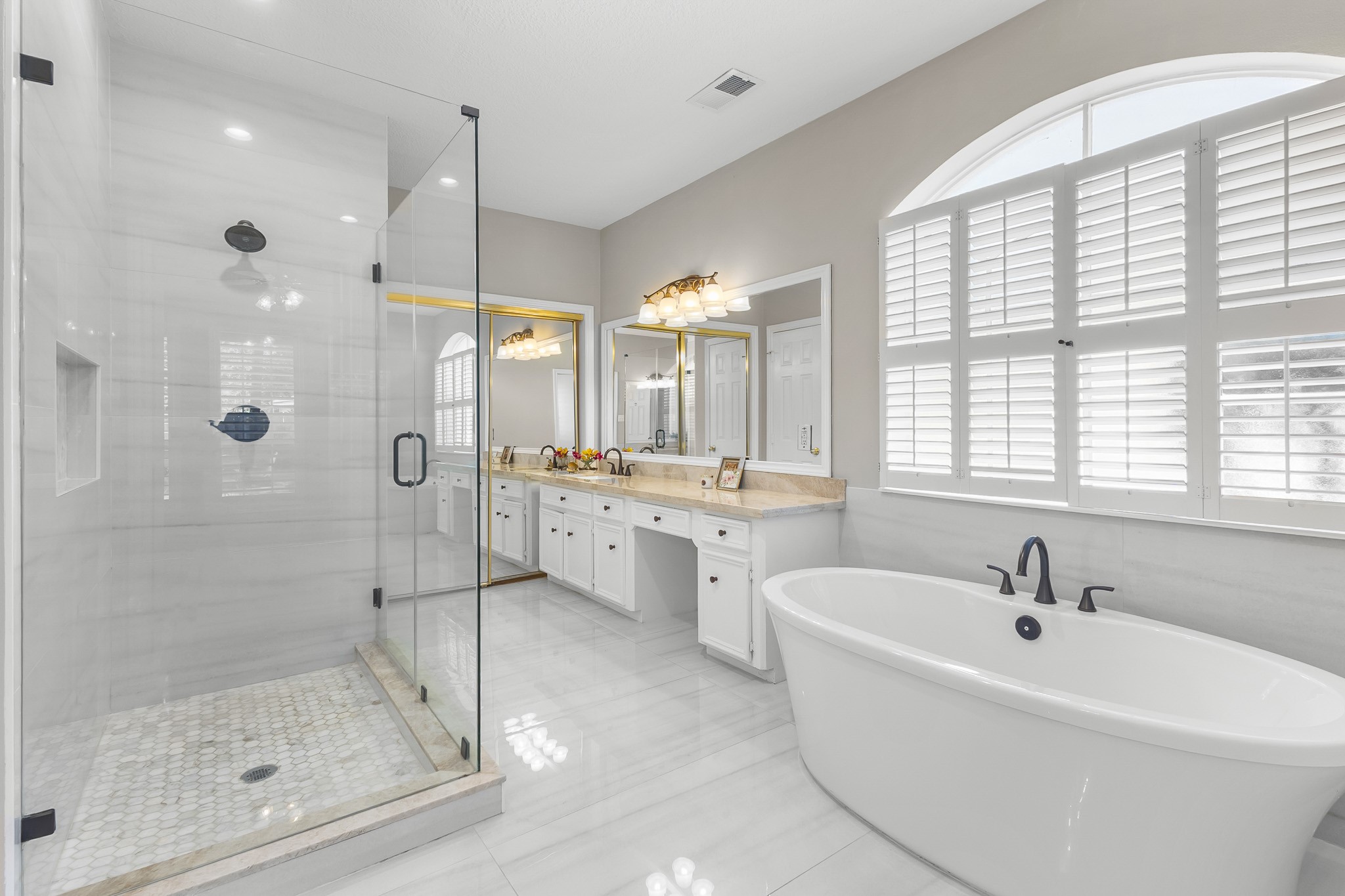 A well-appointed primary bath features a soaking tub, glass-enclosed shower, dual vanities and an expansive arched window.