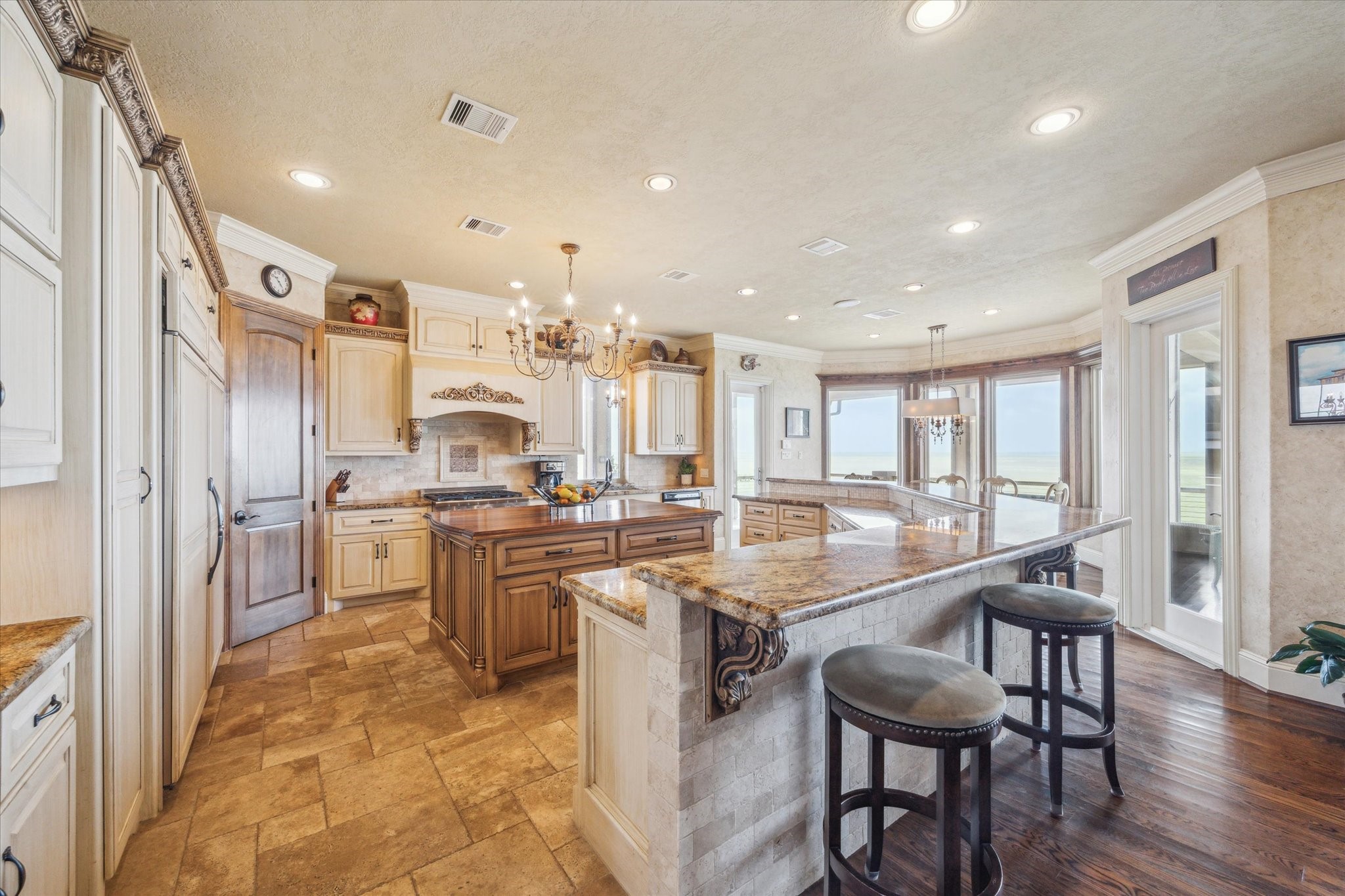 The beautiful kitchen features a large central island, rich wood cabinetry, and warm granite countertops. High-end appliances. Ornate details, custom cabinetry, and expansive bayfront windows complete the timeless Mediterranean design
