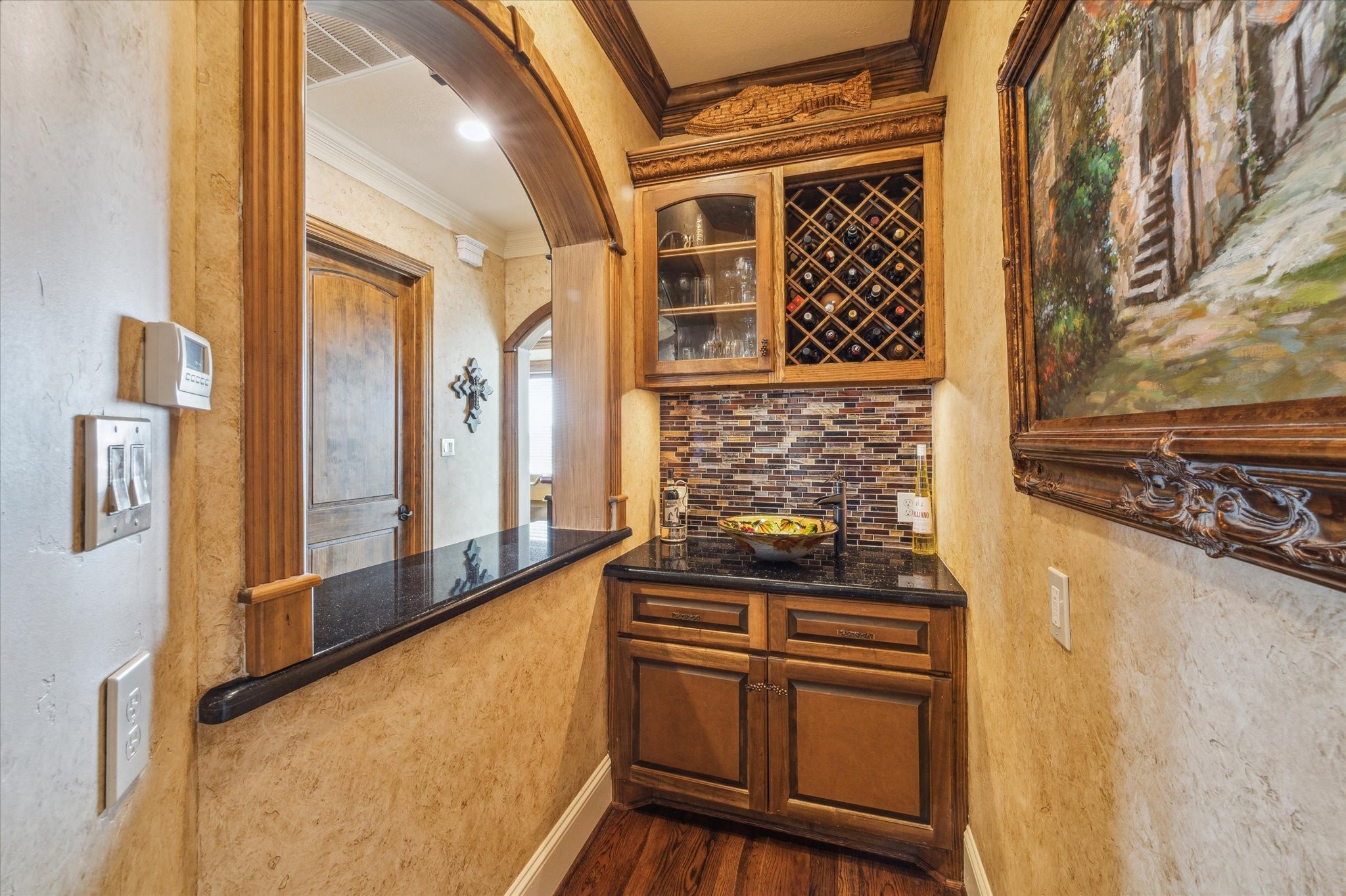 This bold and unique wet bar features custom wood cabinetry, a built-in wine rack, striking granite countertops, and a tile backsplash. An arched pass-through enhances the connection to the surrounding living spaceperfect for entertaining