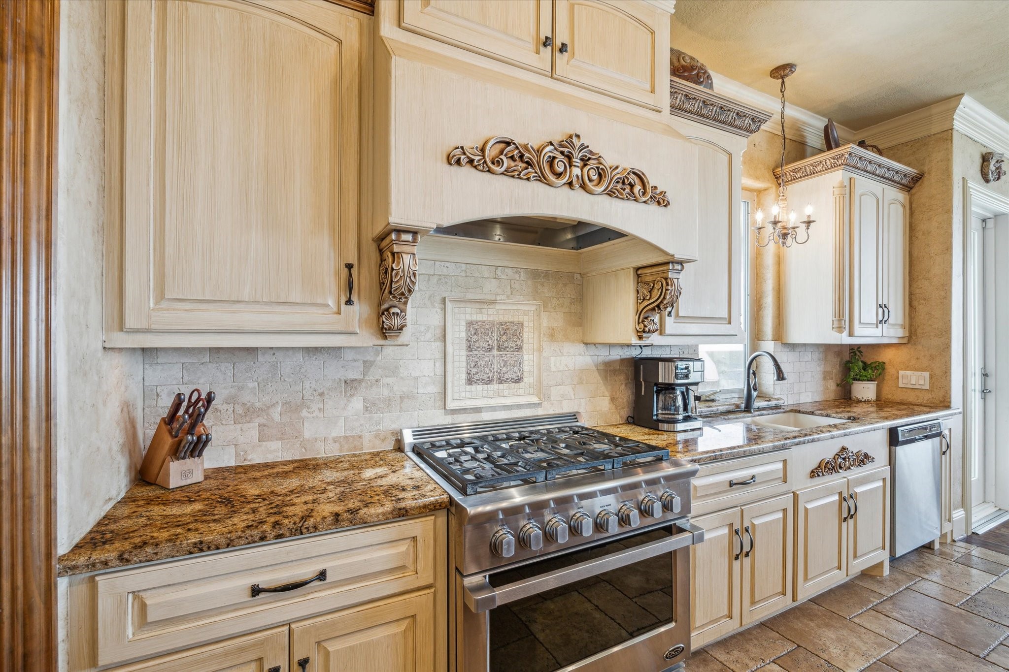 At the heart of the space is a DCS 6-burner gas range and convection oven, set against a backdrop of warm, tumbled stone backsplash tiles and custom mosaic tiled design. Above the range, a stunning custom wood range hood takes center stage, adorned with intricate carved scrollwork and corbels that echo Old World design influences.