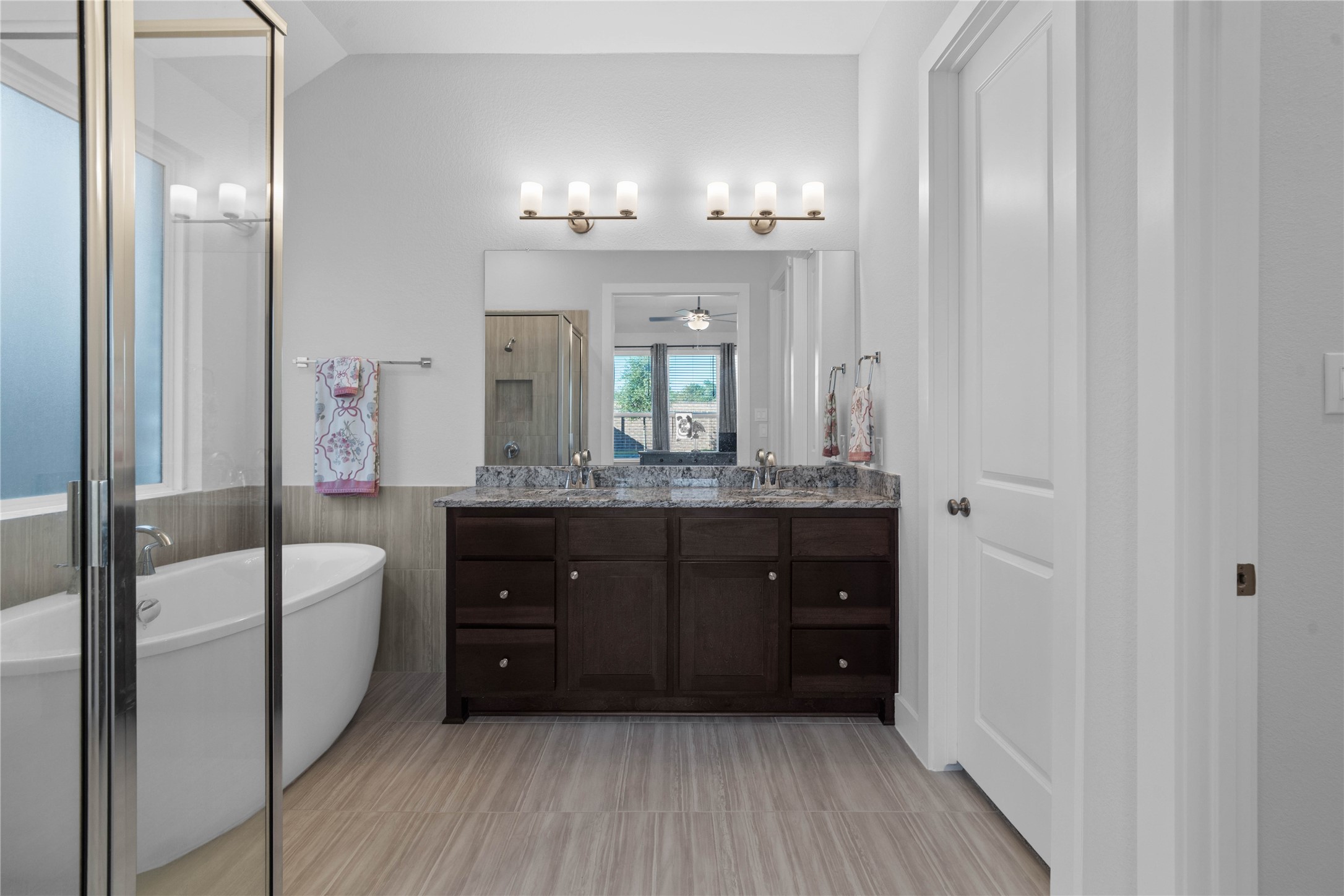 The luxurious primary en-suite bathroom is a true spa-like oasis, showcasing an extended dual-sink granite vanity, rich dark wood cabinetry, premium lighting fixtures, a modern freestanding soaking tub, and a separate glass-enclosed walk-in shower.