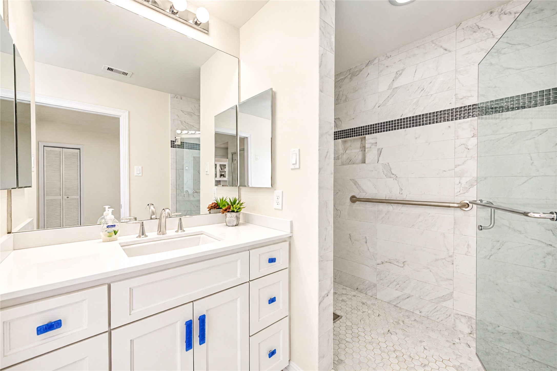 A bright vanity with a light countertop faces a frameless walk-in shower finished with a mosaic accent band.