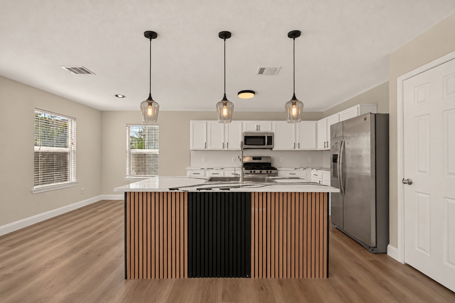 A substantial central island anchors the kitchen beneath pendant lighting, while abundant white cabinetry lines the walls to keep storage close at hand. Clean finishes and ample work surfaces give the renovated space a fresh, contemporary look.