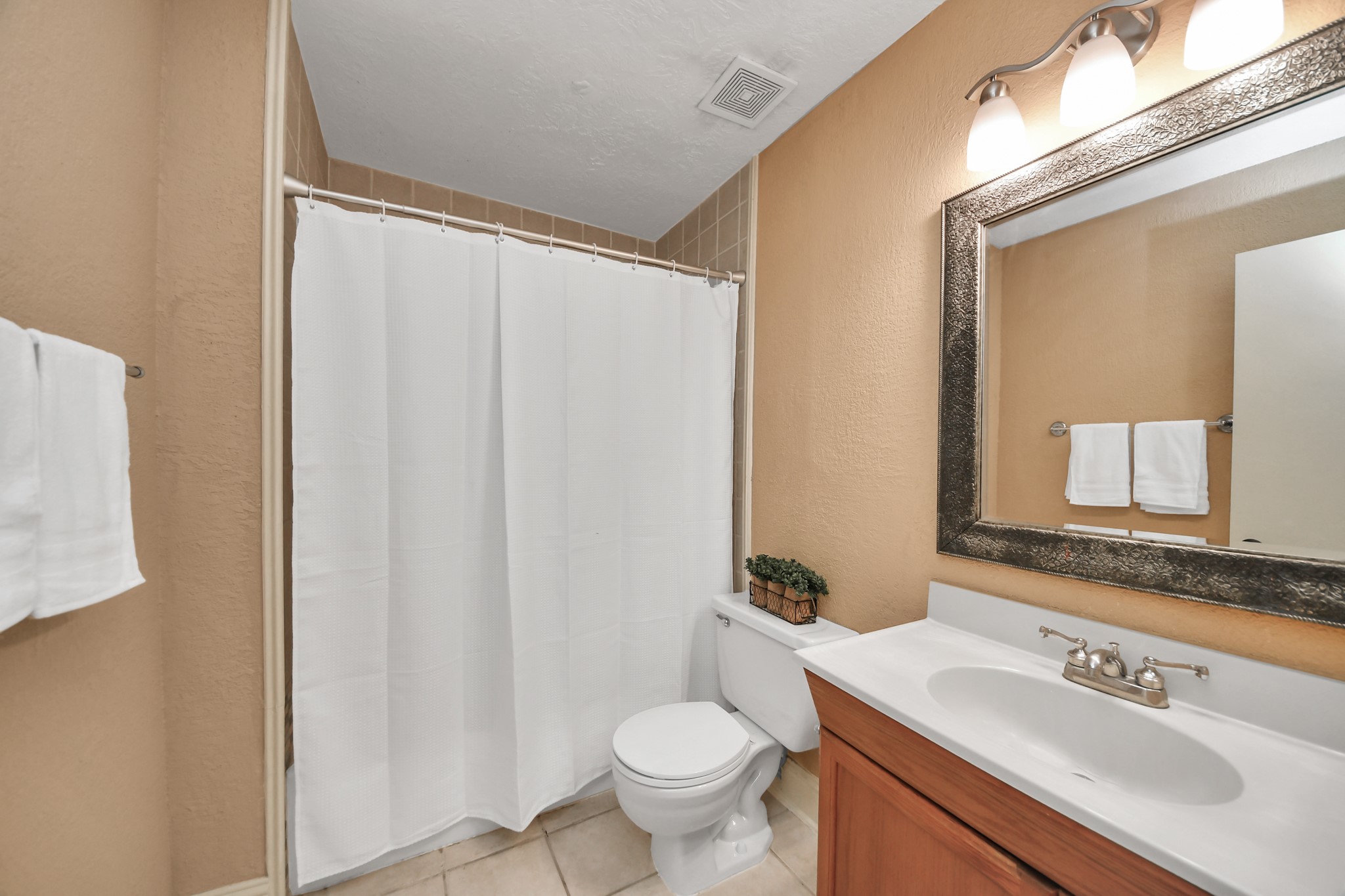 The upper-level full bathroom offers a welcoming combination of comfort and practicality. Warm neutral walls complement the wood-tone vanity, while a spacious countertop, decorative framed mirror, and multi-light vanity fixture add character to the space.