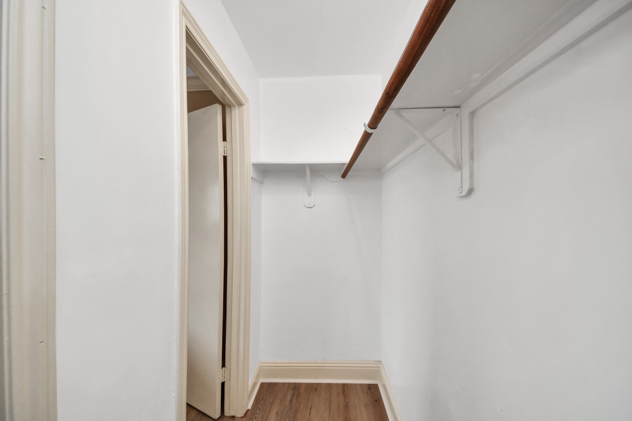 White walls provide a clean, bright appearance, while the built-in hanging rods and overhead shelving offer room for clothing, shoes, and accessories. The wood-look flooring continues into the closet, creating a cohesive transition from the bedroom.