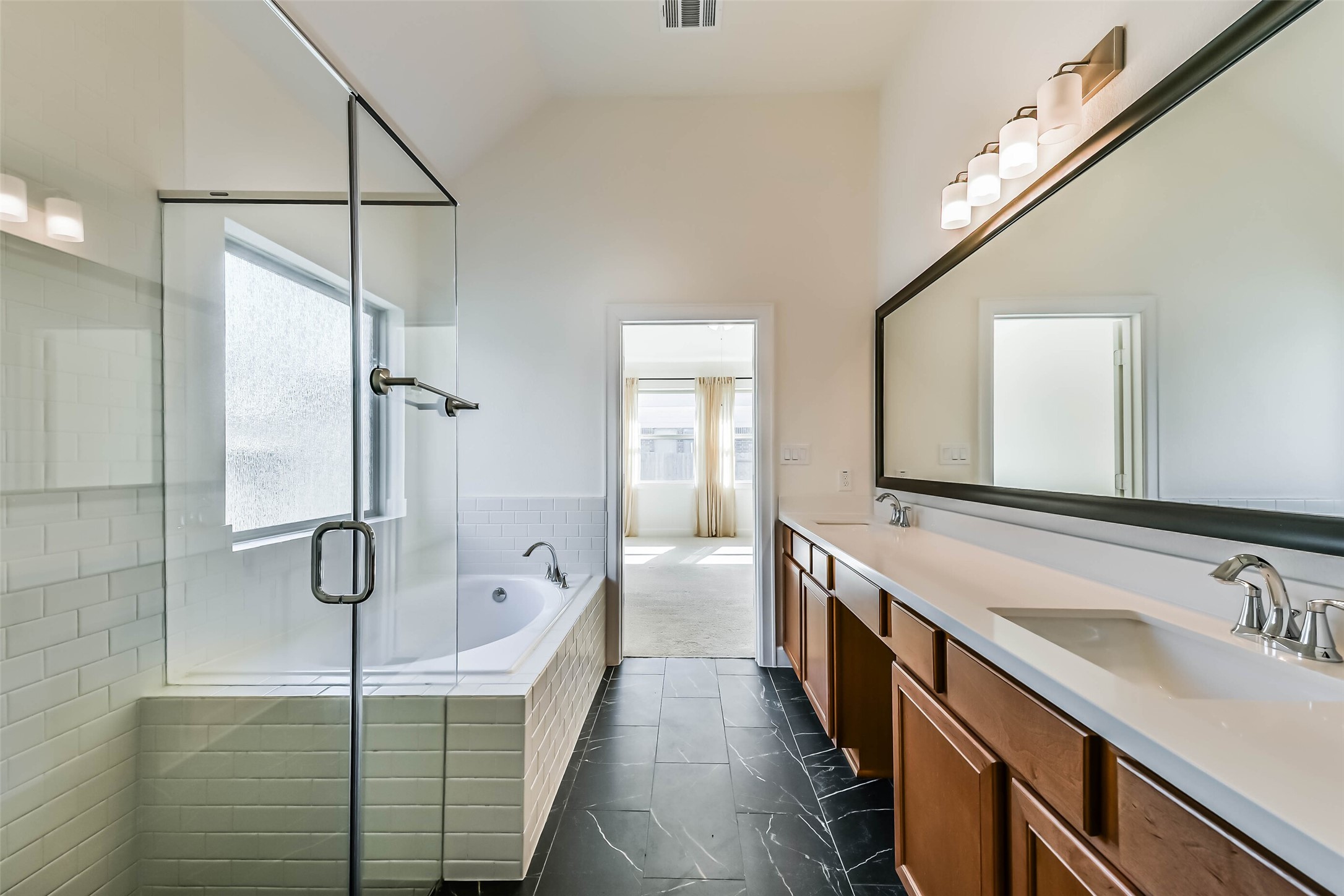 Spacious primary bathroom featuring a luxurious glass-enclosed shower, a relaxing soaking tub, dual sinks with elegant fixtures, and sleek black tile flooring.
