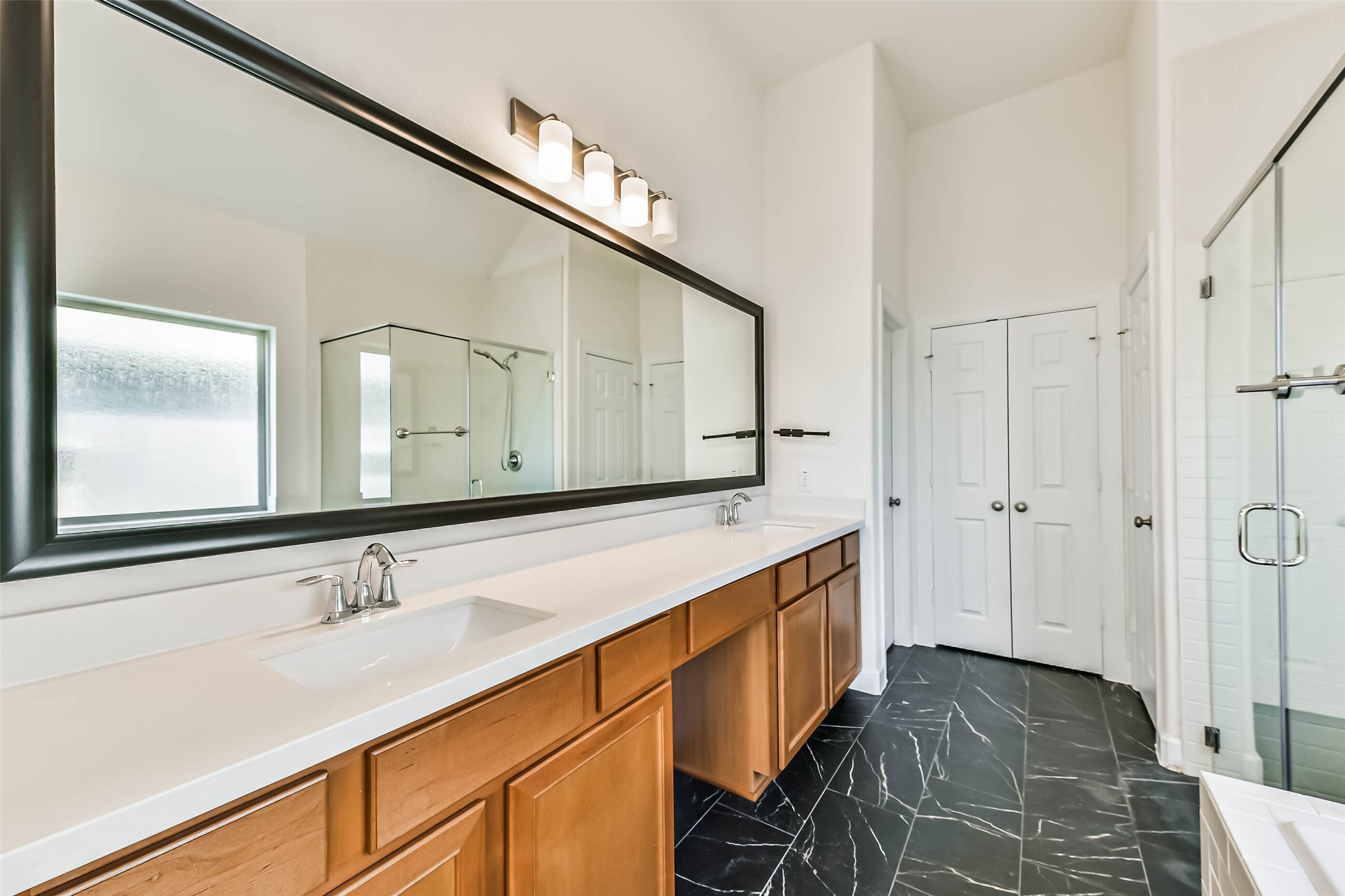 Spacious primary bathroom featuring a double vanity with sleek countertops, a large mirror, elegant black-tiled flooring, and a glass-enclosed shower for a luxurious touch.