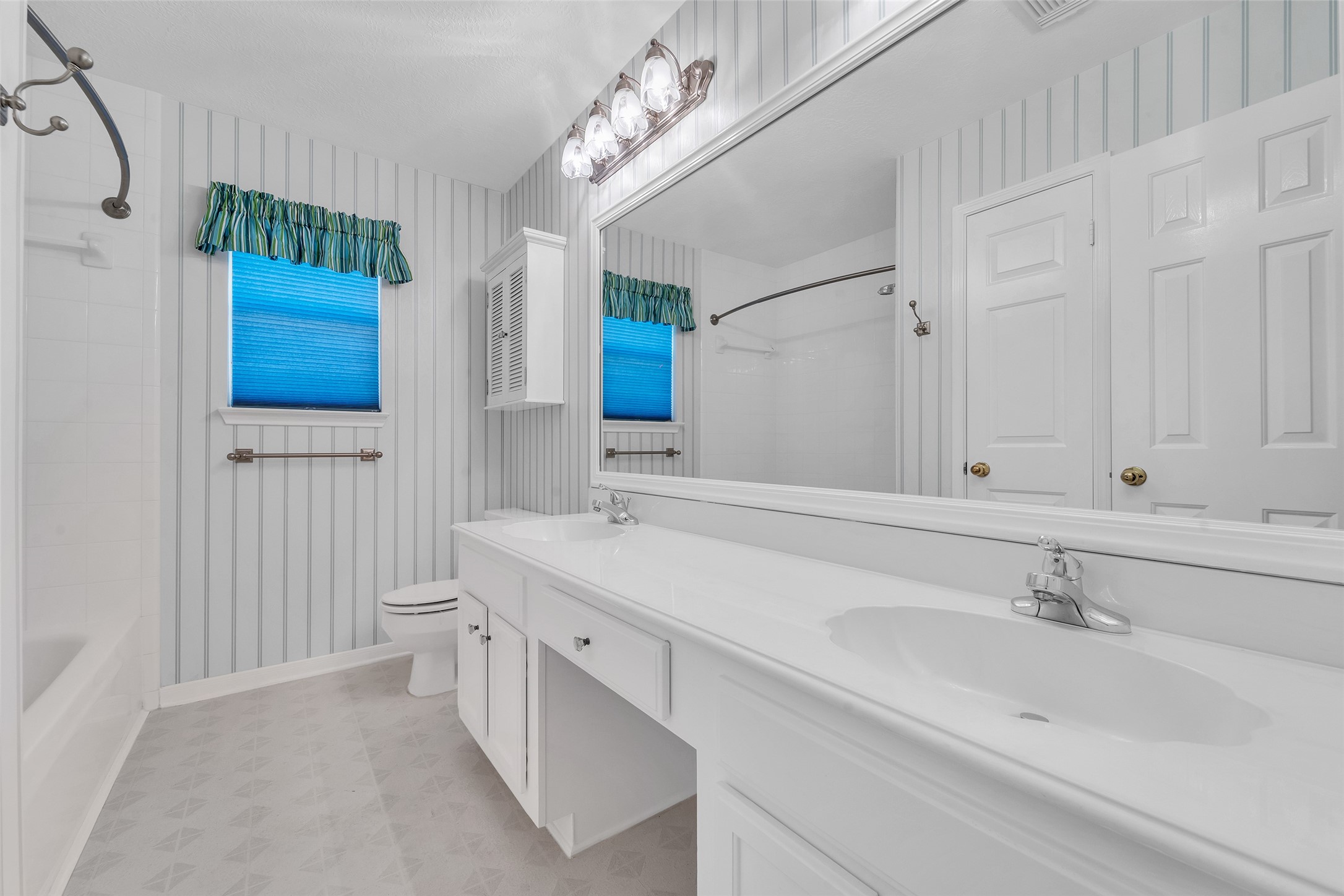 Bright and spacious bathroom featuring a large vanity with double sinks, elegant striped wallpaper, and a combination tub and shower.