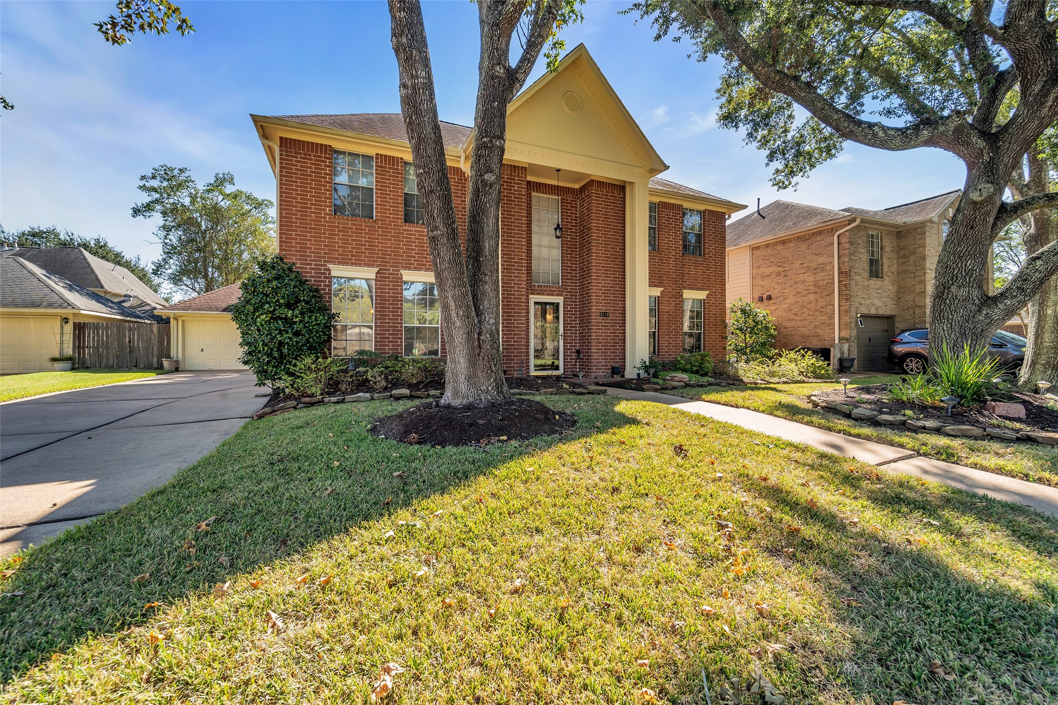Charming two-story brick home with a welcoming entrance, mature trees, and a spacious driveway leading to a detached garage.