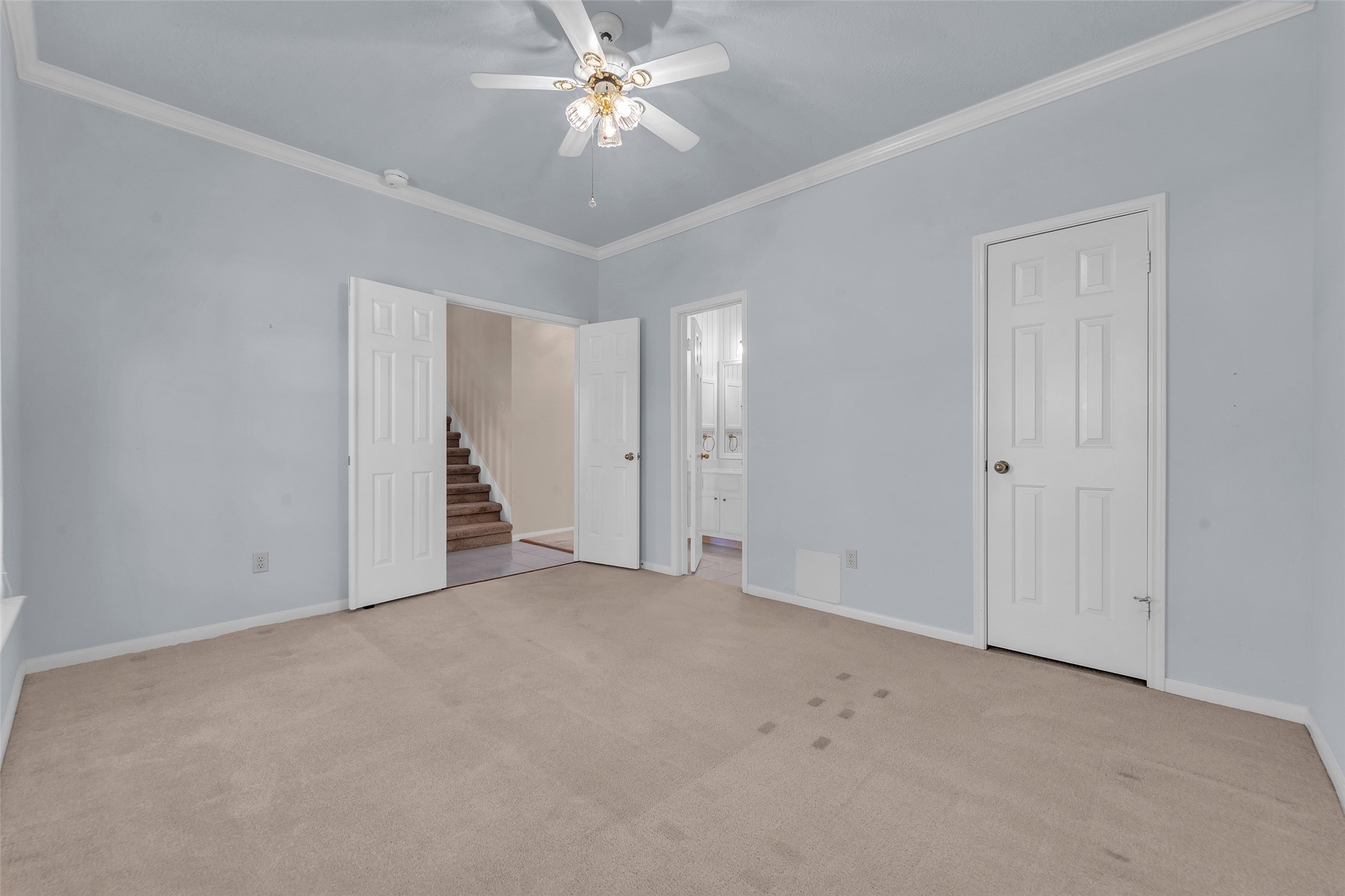 Spacious bedroom with soft blue walls, crown molding, and a ceiling fan, offering access to a bathroom and nearby stairway.