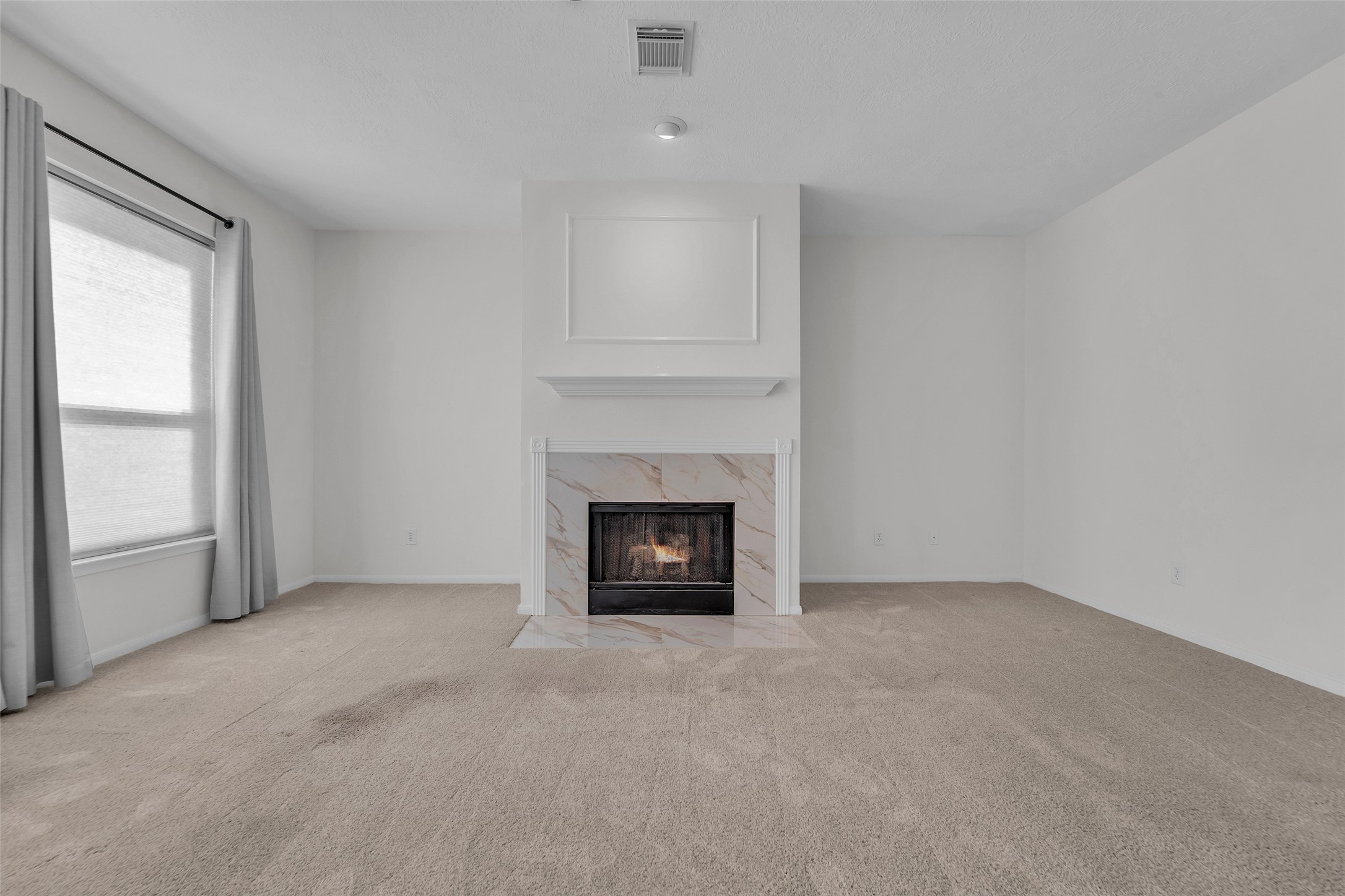 Spacious living room featuring a cozy fireplace with elegant marble detailing, complemented by soft carpet and abundant natural light from a large window.