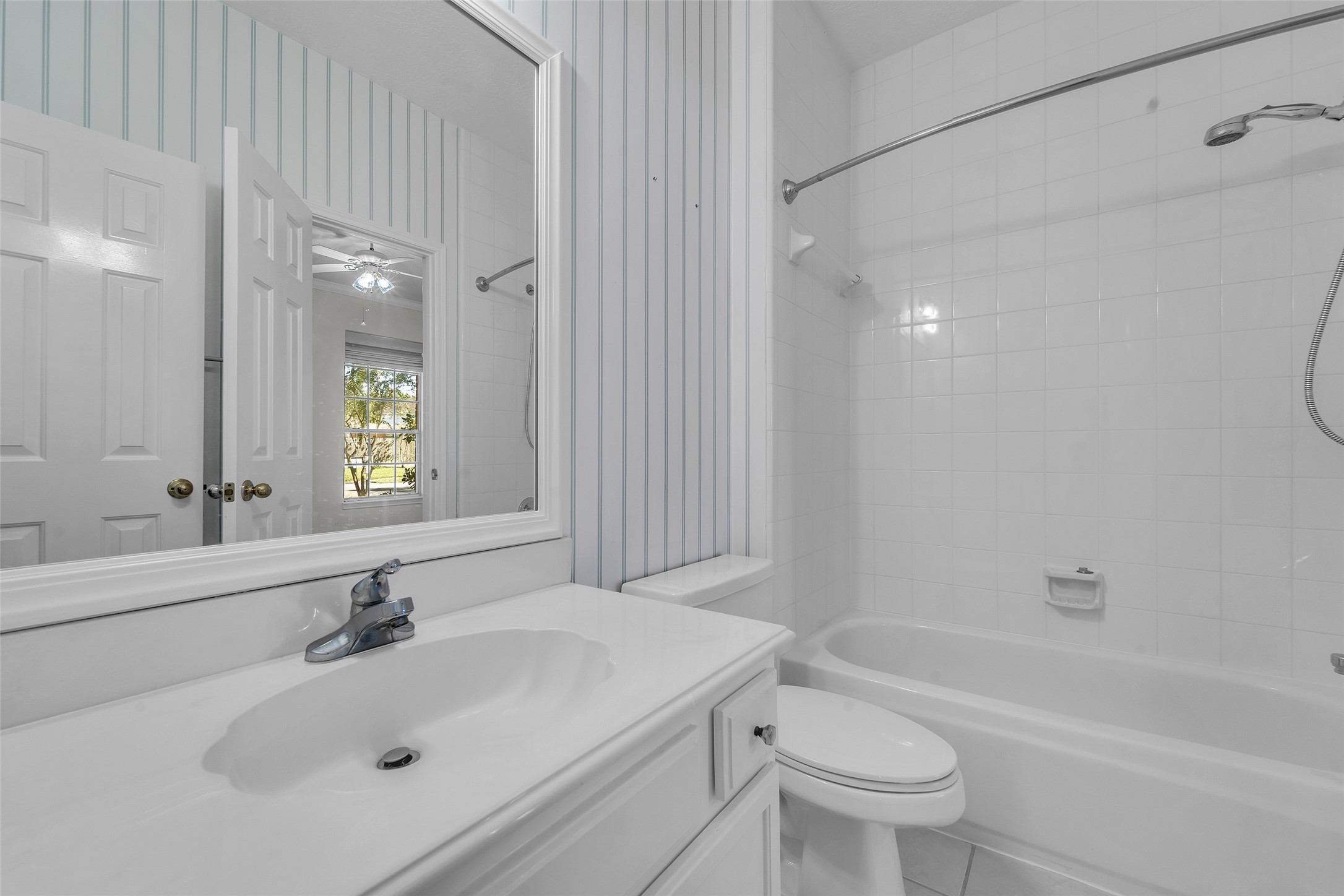 Bright and clean bathroom featuring a classic white tile bathtub, modern sink, and a large mirror with elegant vertical striped wallpaper for a touch of charm.