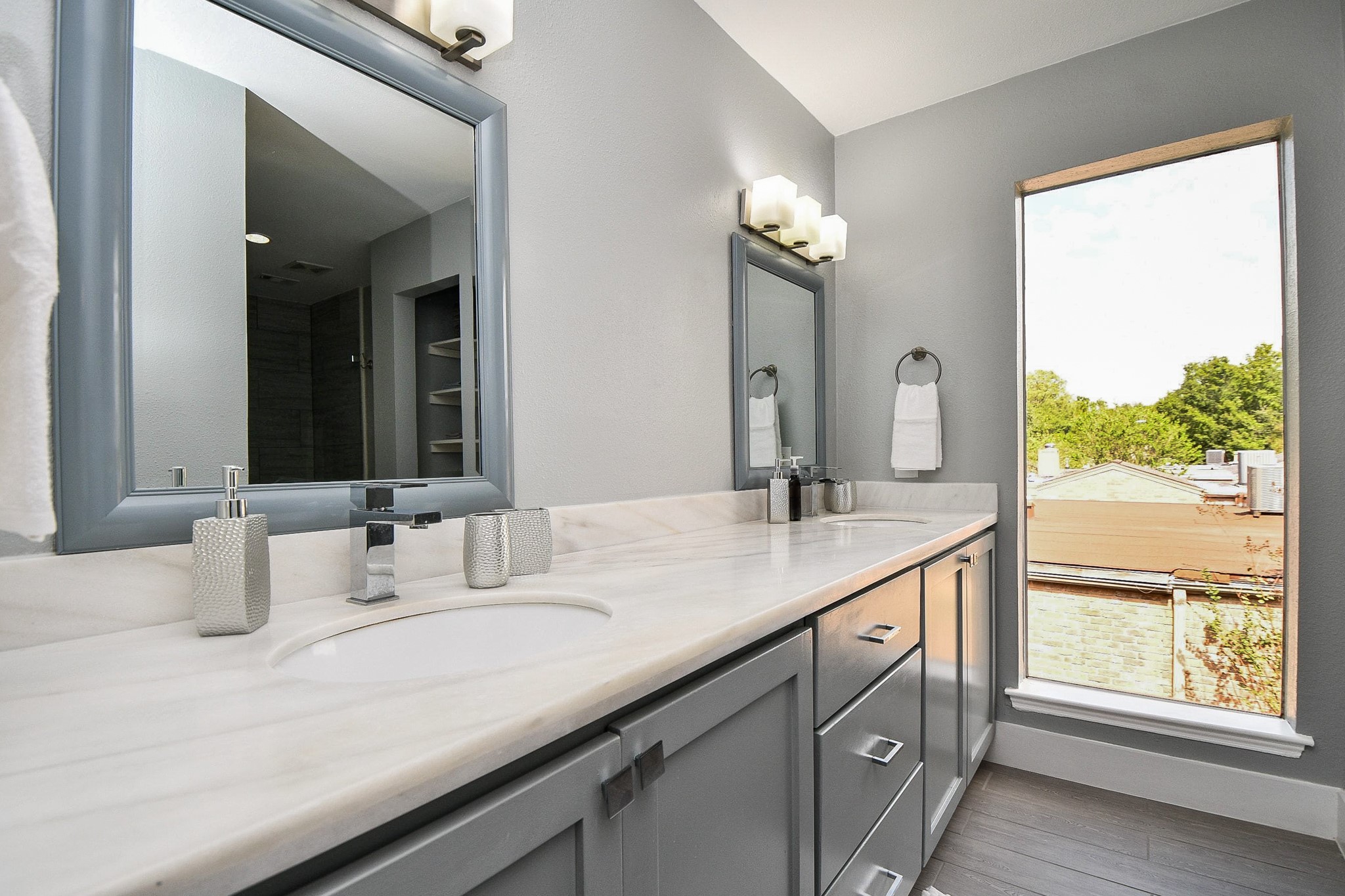 The updated primary bath has a long vanity with gray cabinetry, stone-look counters, and a tall window for natural light.