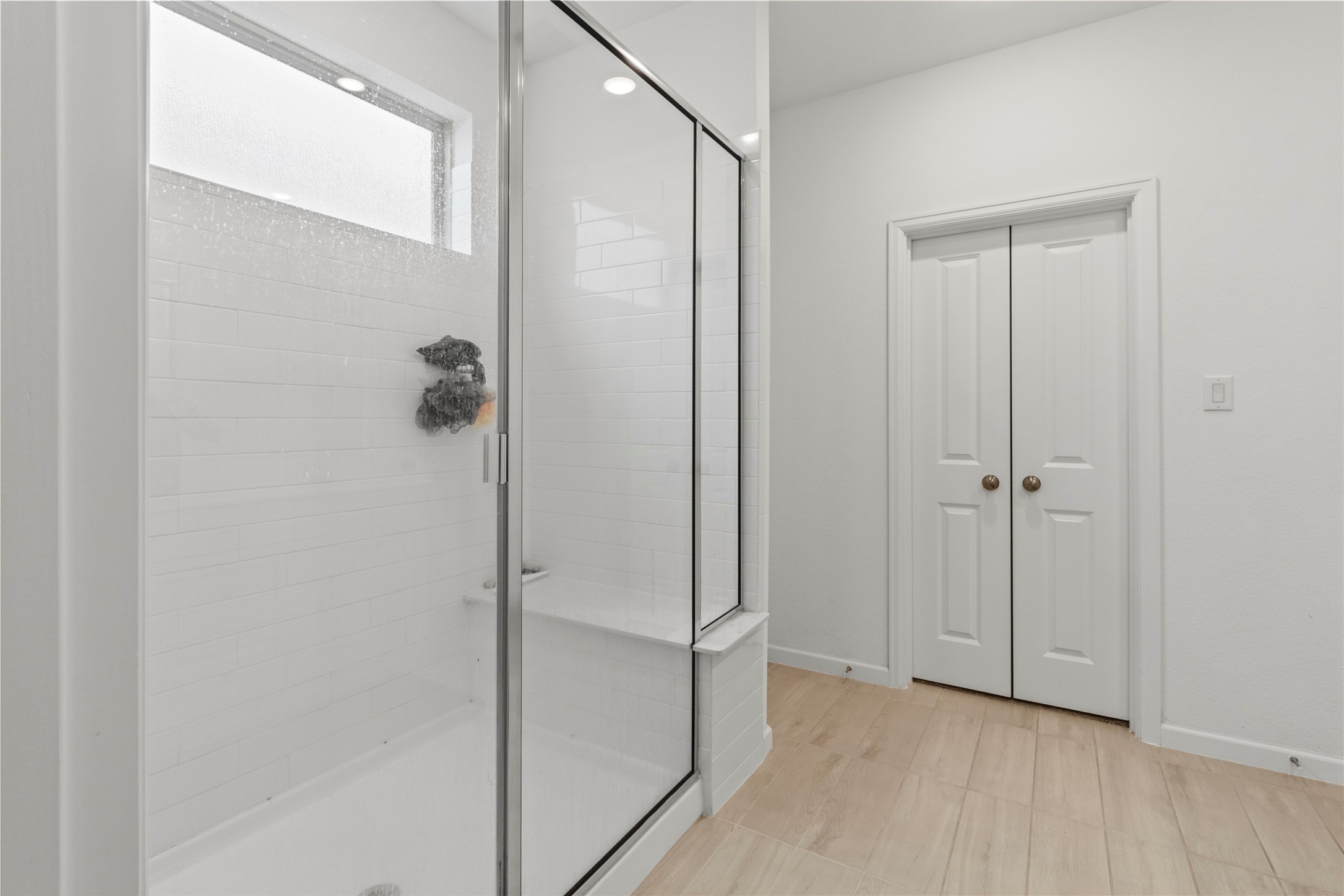 Sleek and modern bathroom off the primary with elegant white tiles, complemented by stylish wood-look flooring and convenient double doors for storage.