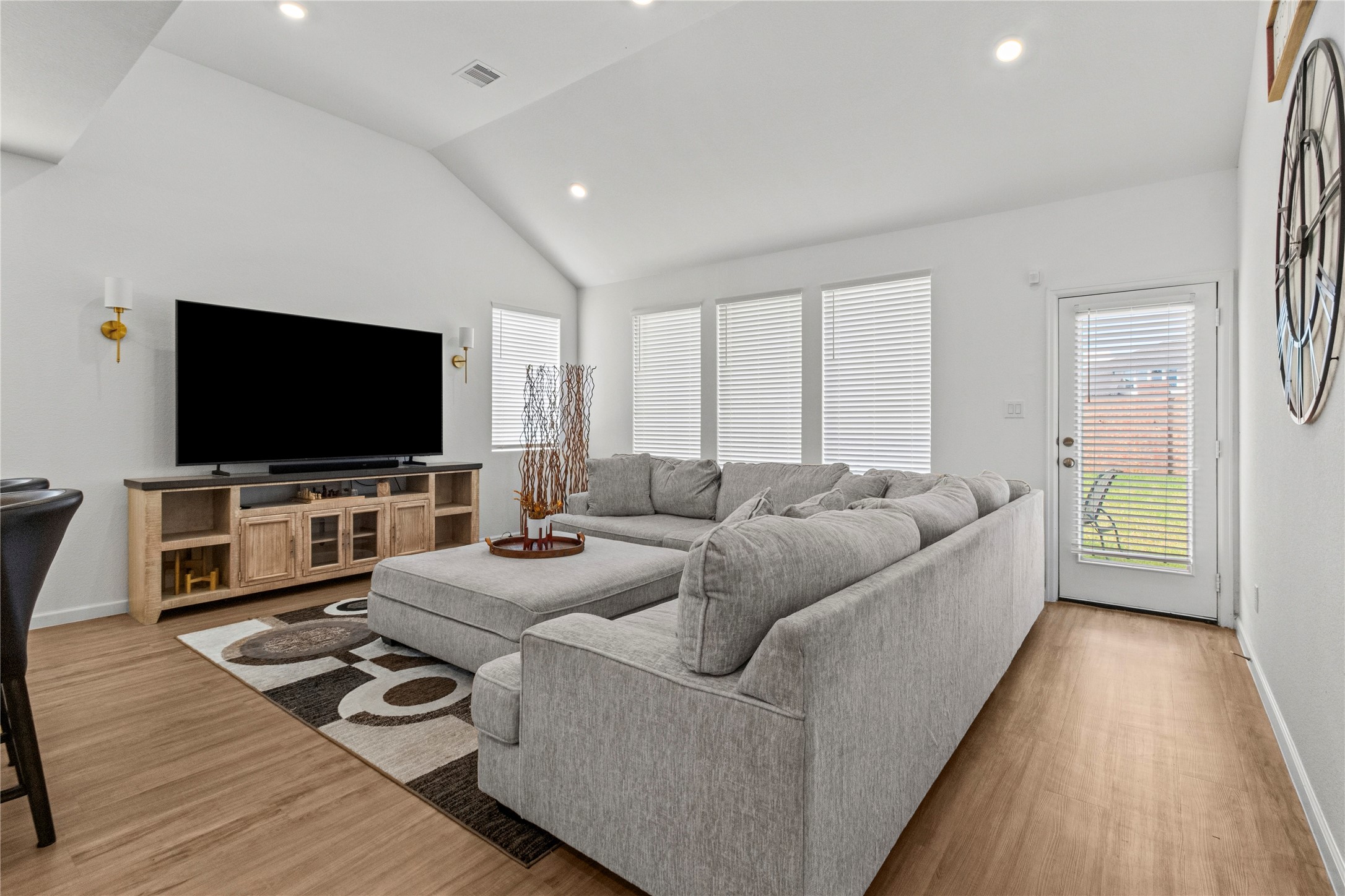 Bright and inviting living area with vaulted ceilings features a spacious sectional sofa, large windows with blinds for natural light, and a modern entertainment setup. A door leads to a backyard space, enhancing indoor-outdoor living.
