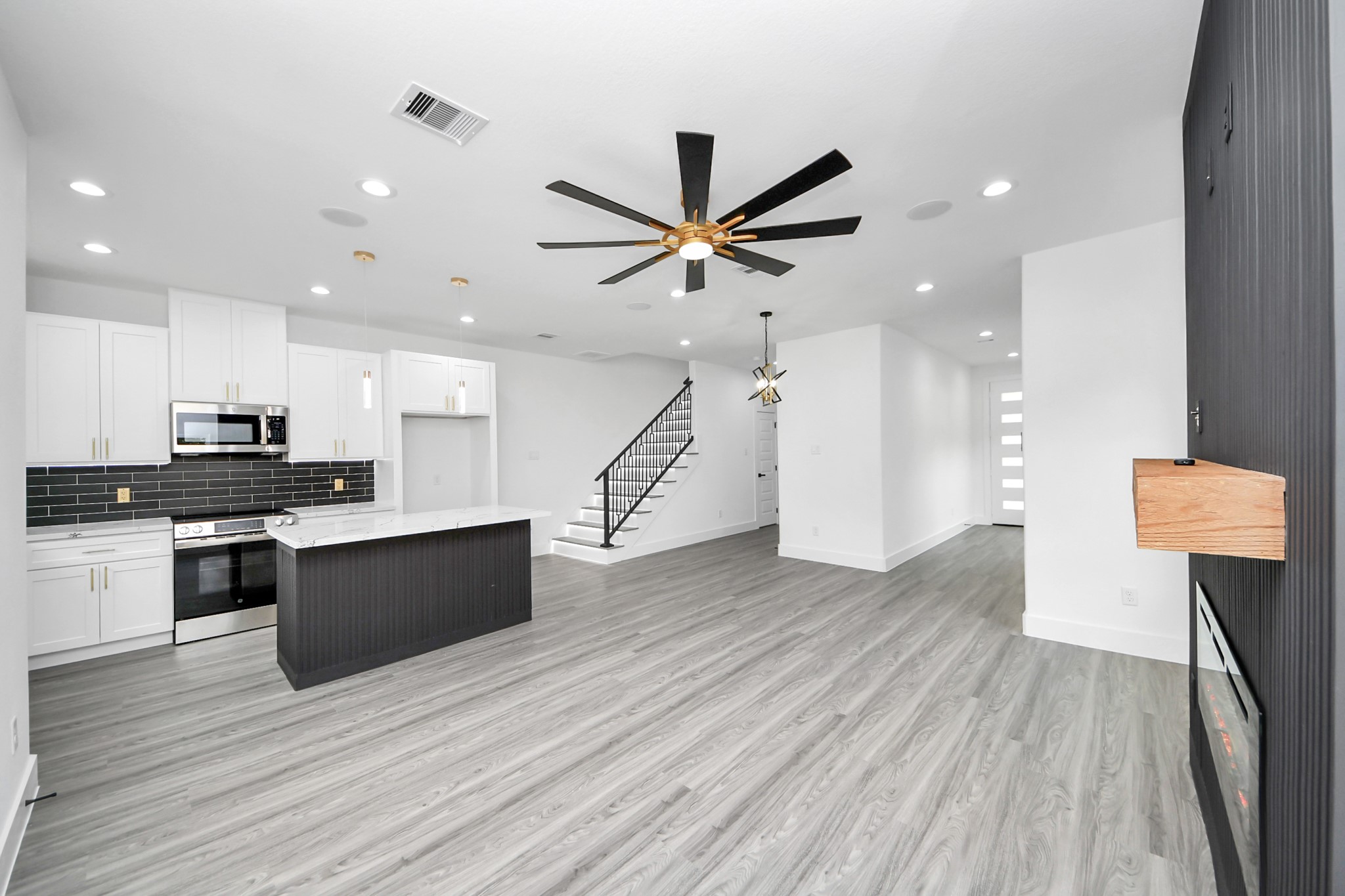 Modern open-concept living area featuring sleek gray flooring, a stylish kitchen with a large island, and contemporary black backsplash. A striking ceiling fan and recessed lighting enhance the space, complemented by a chic staircase leading to the upper level.