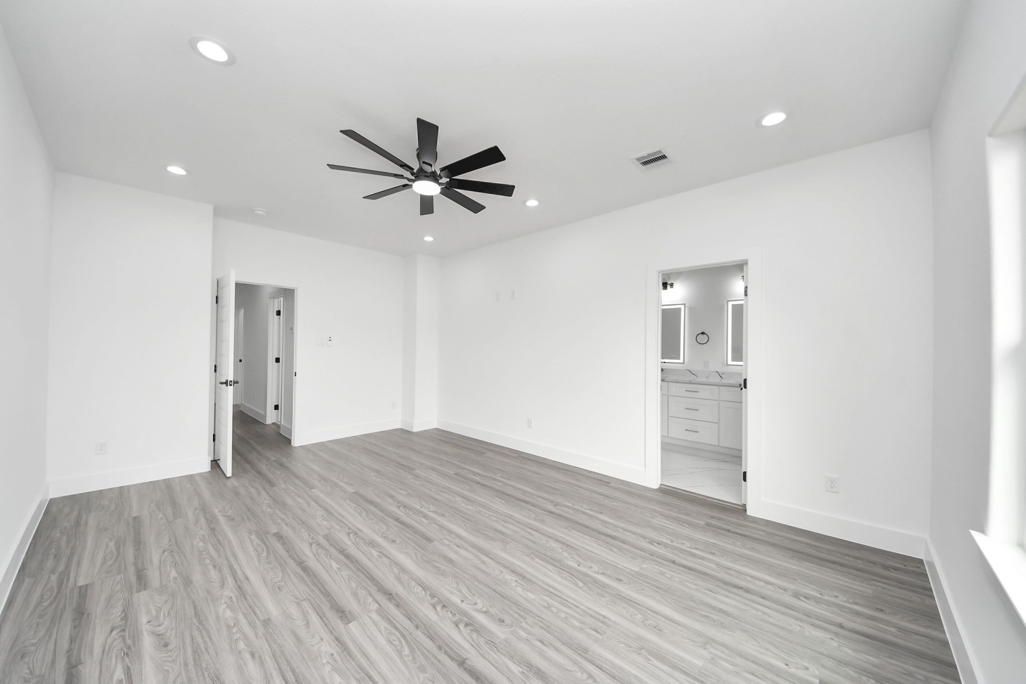 Spacious, modern room featuring sleek wood flooring, recessed lighting, and a ceiling fan, with direct access to a bright, stylish bathroom.