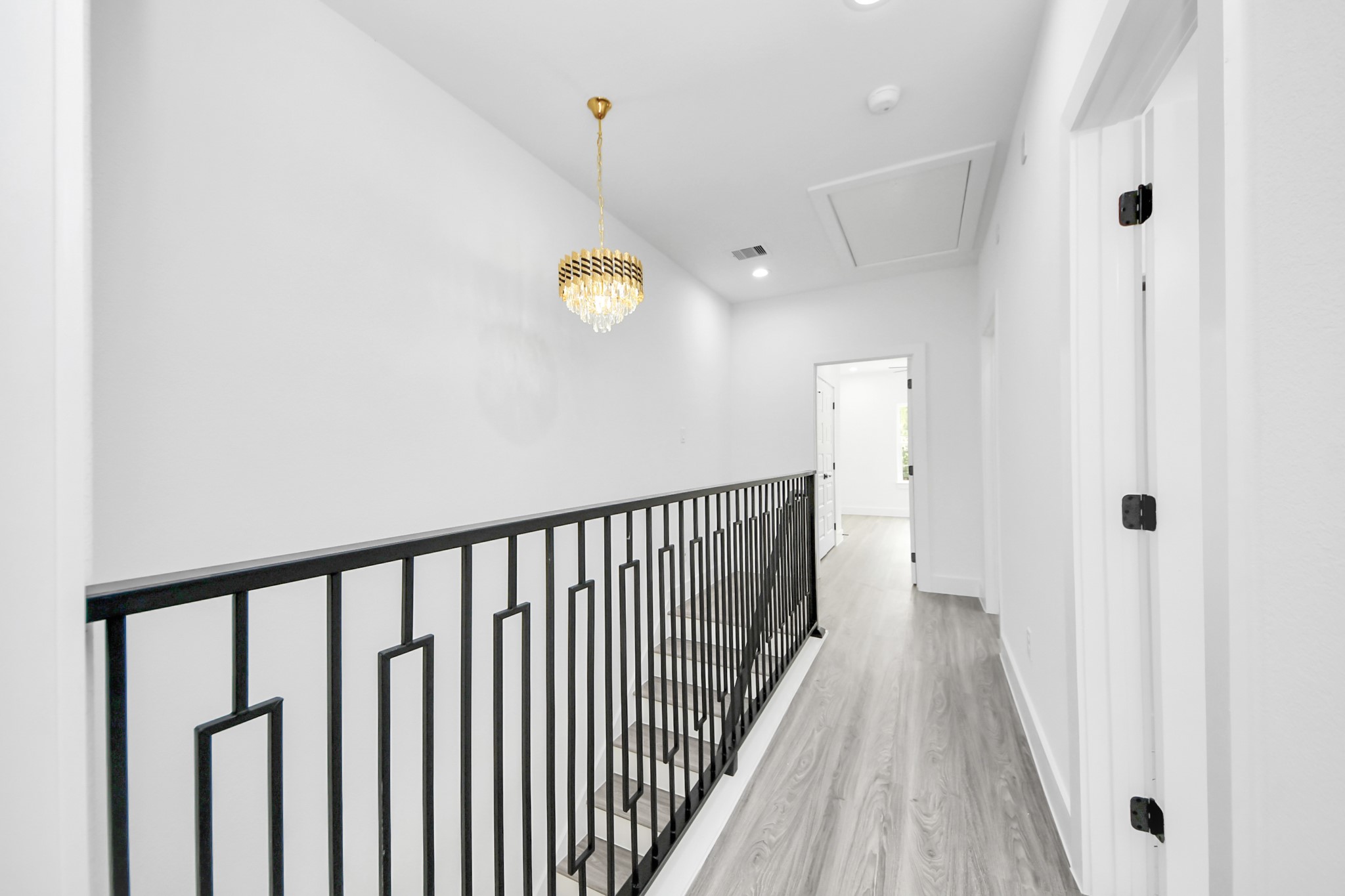 Elegant hallway featuring modern railing, a stylish chandelier, and sleek wood flooring, leading to bright rooms.