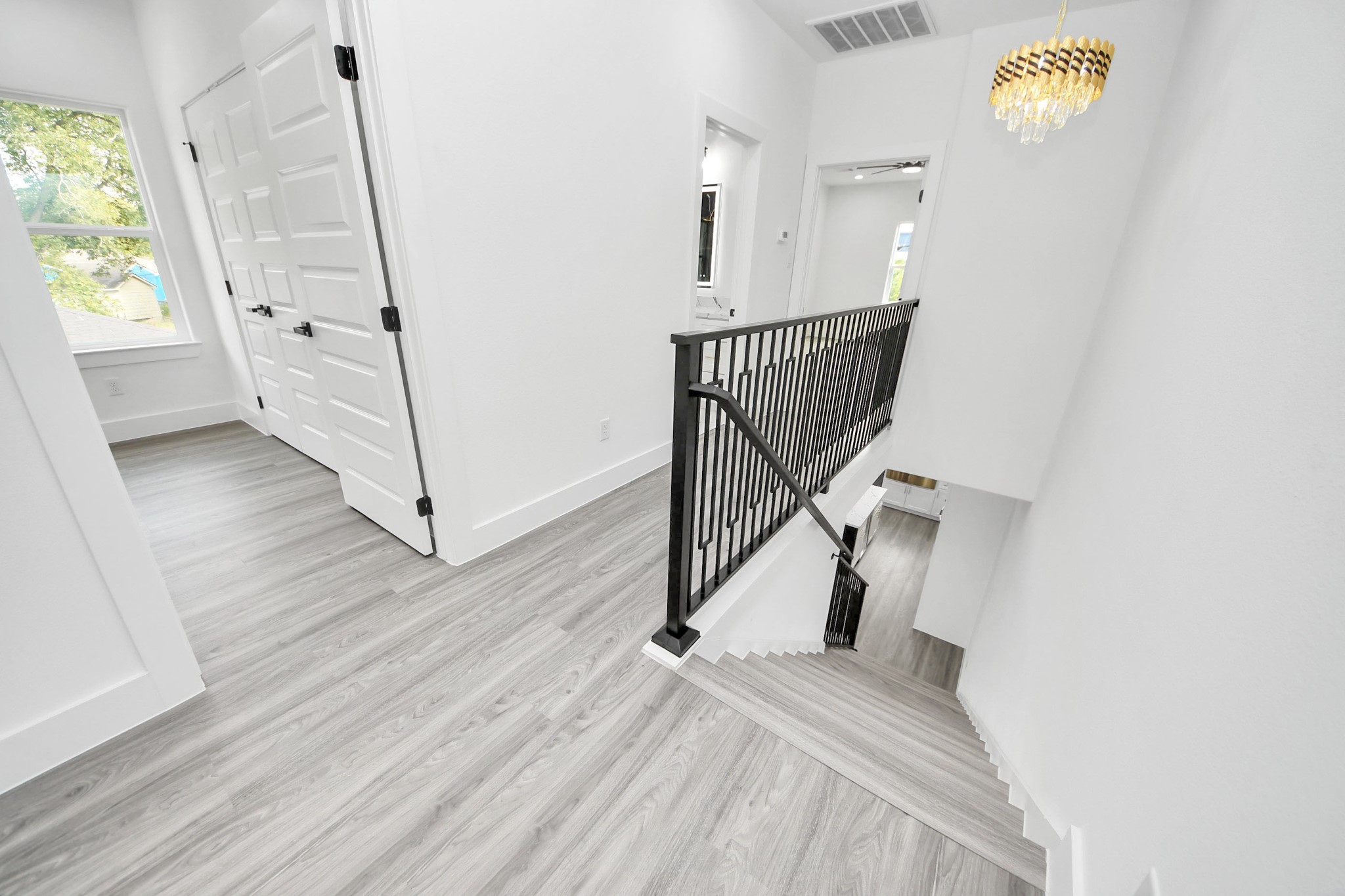 Bright and modern hallway with sleek wood flooring, elegant black railing, and a chic chandelier accentuating the airy space.
