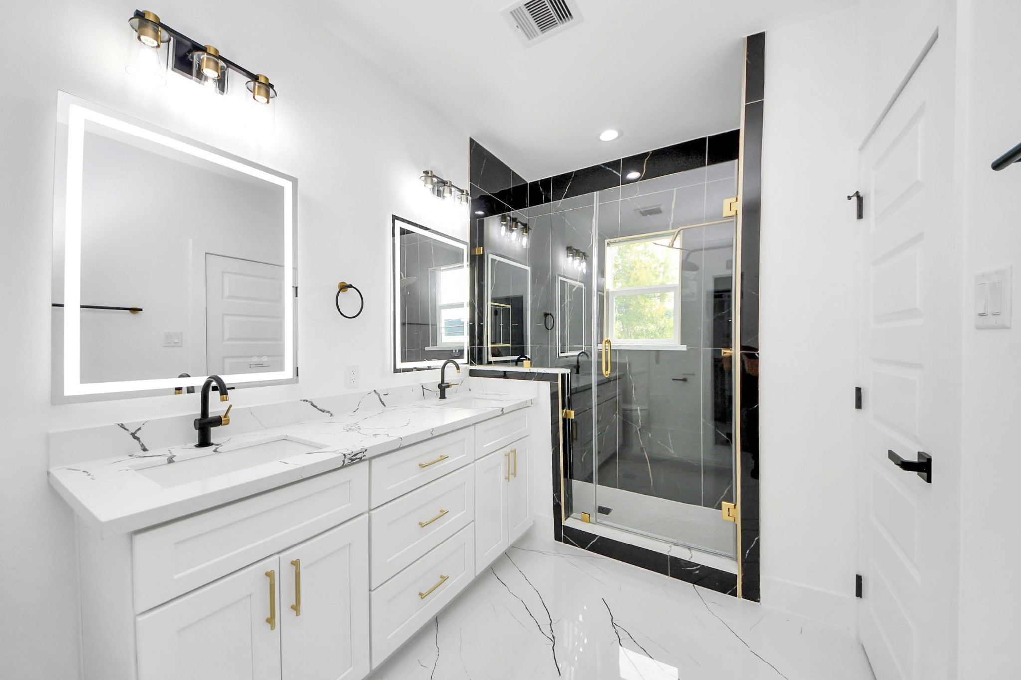Luxurious bathroom featuring a spacious dual-sink vanity with elegant marble countertops, sleek black fixtures, and a stylish glass-enclosed shower accented with gold hardware.