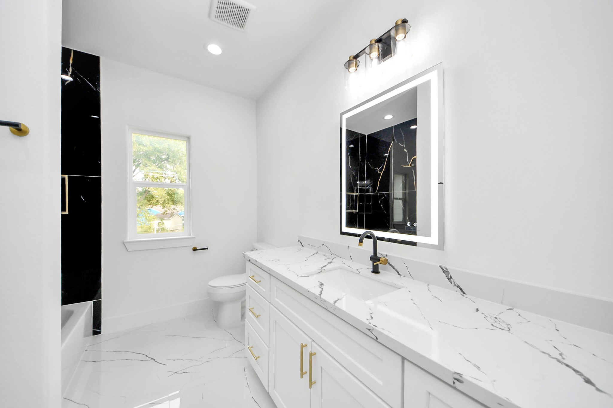 Elegant bathroom featuring a sleek, marble-topped vanity with modern fixtures, complemented by a stylish illuminated mirror and luxurious black accent wall.