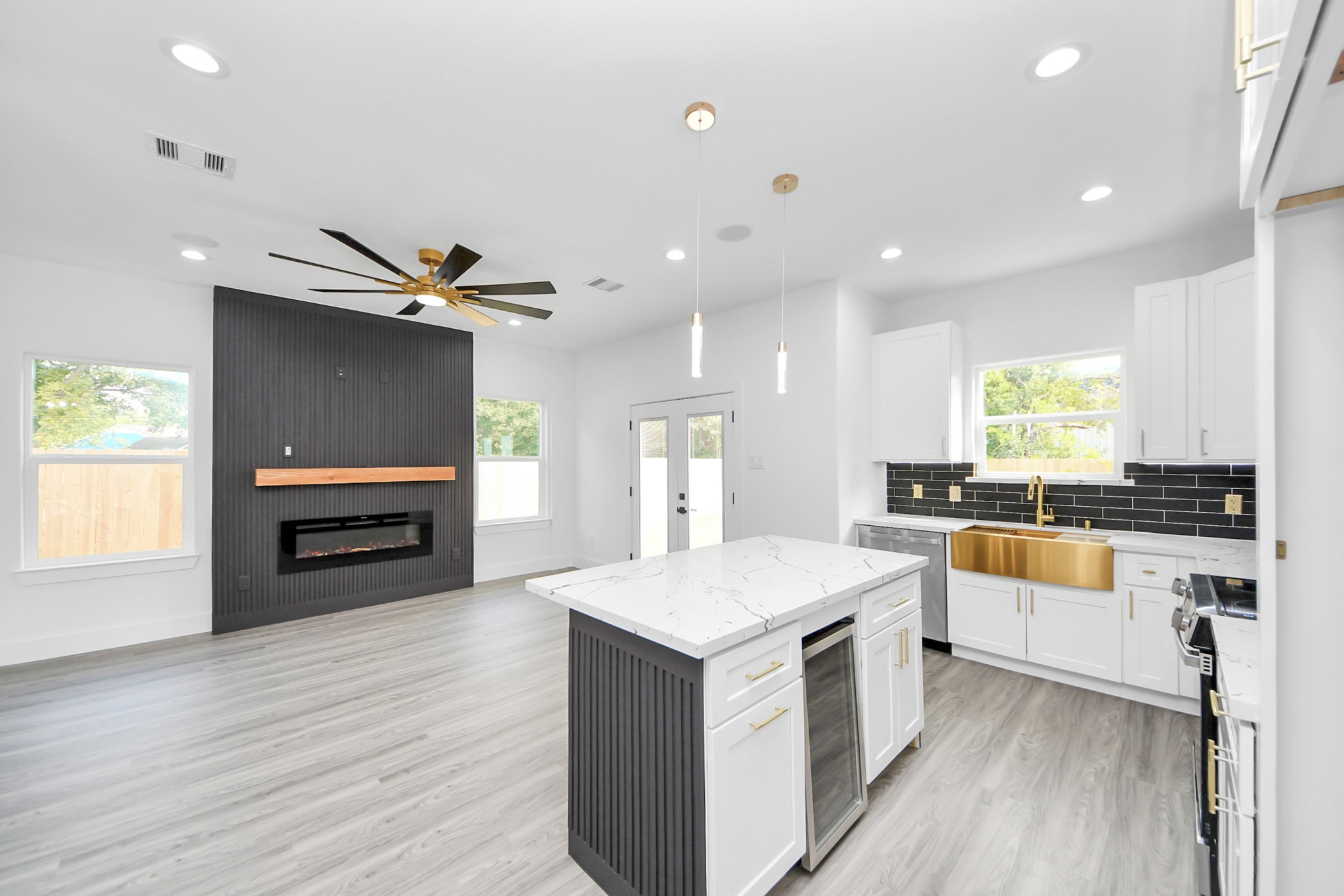 Experience modern elegance in this open-concept living space featuring a sleek kitchen with a large island, gold accents, and a striking black backsplash. The adjacent living area offers a contemporary fireplace and stylish ceiling fan, all enhanced by abundant natural light.