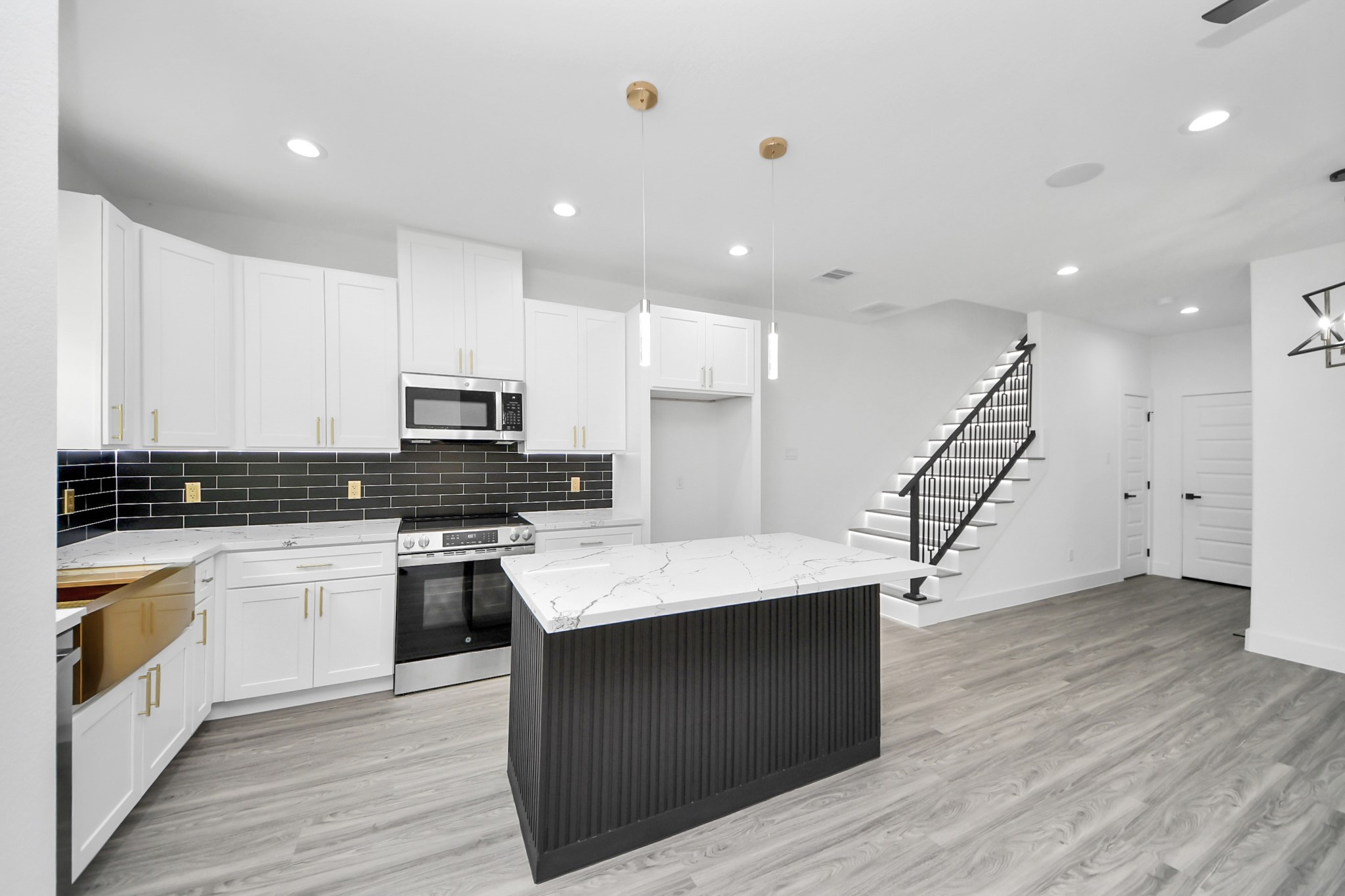 Sleek modern kitchen with white cabinetry, marble countertops, and a stylish black tile backsplash. The open layout flows seamlessly into the living area, accented by elegant pendant lighting and a contemporary staircase.