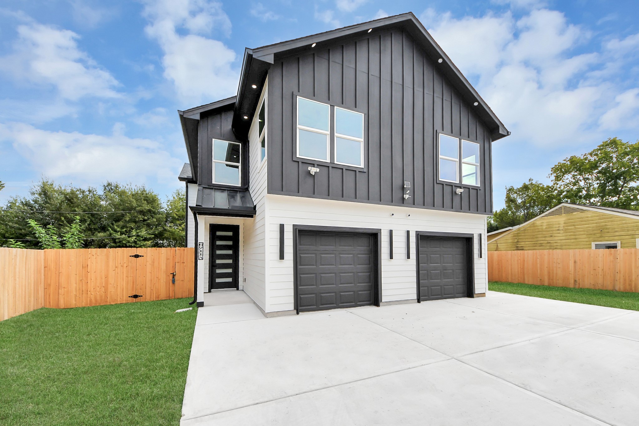 Modern duplex featuring a sleek black facade with two single-car garages, set on a spacious driveway and surrounded by a wooden privacy fence.