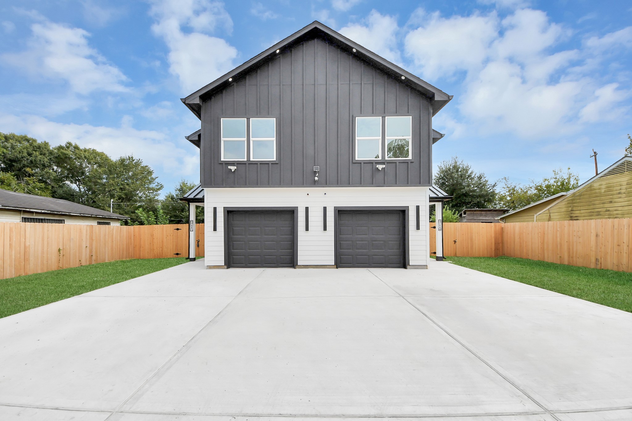 Modern duplex with a sleek dark exterior, featuring two spacious garages and a large concrete driveway. The property is enclosed by a wooden fence, offering a private and serene setting.