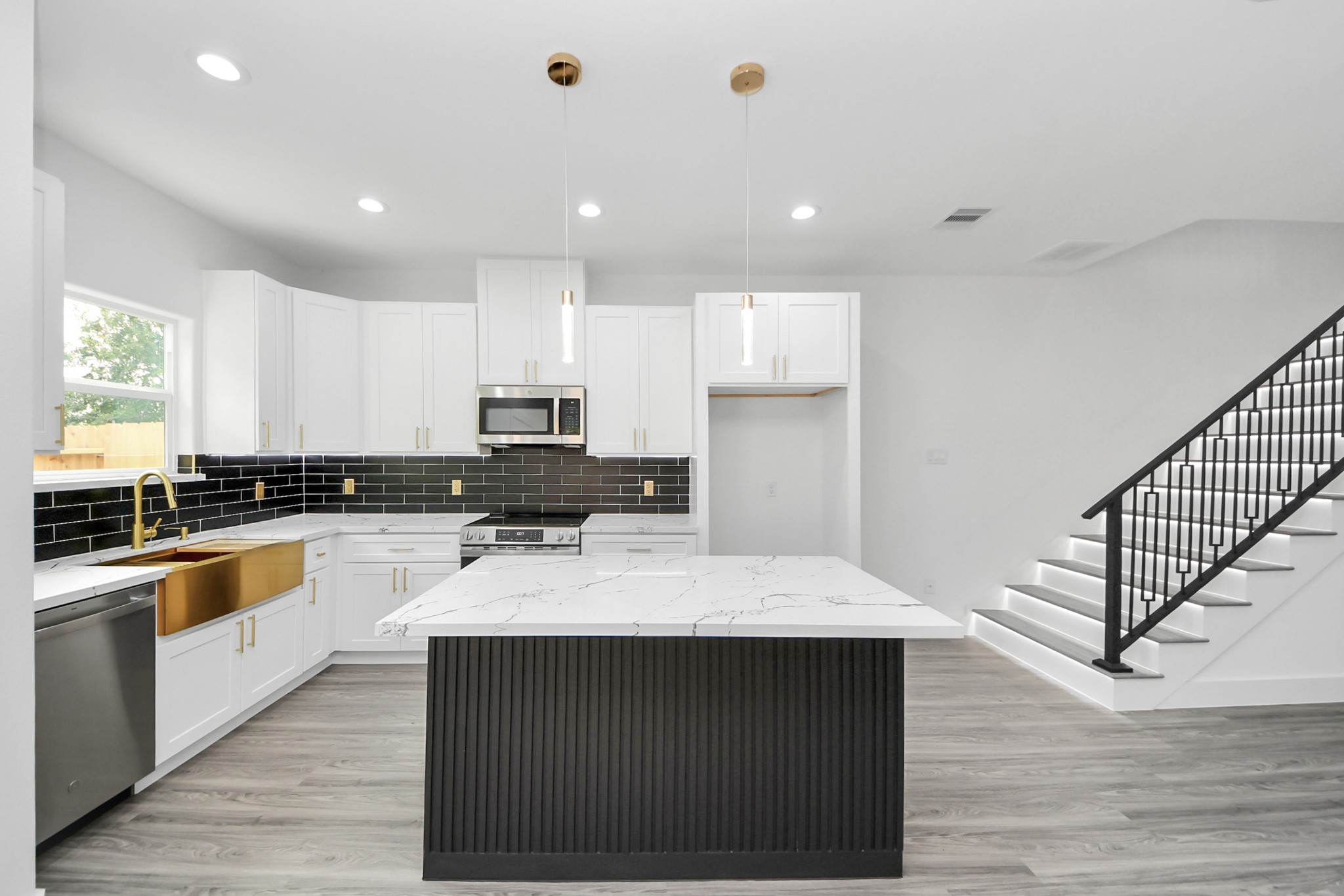 Sleek modern kitchen featuring a large island with marble countertops, gold accents, and stylish black tile backsplash, complemented by elegant pendant lighting and adjacent to a contemporary staircase.